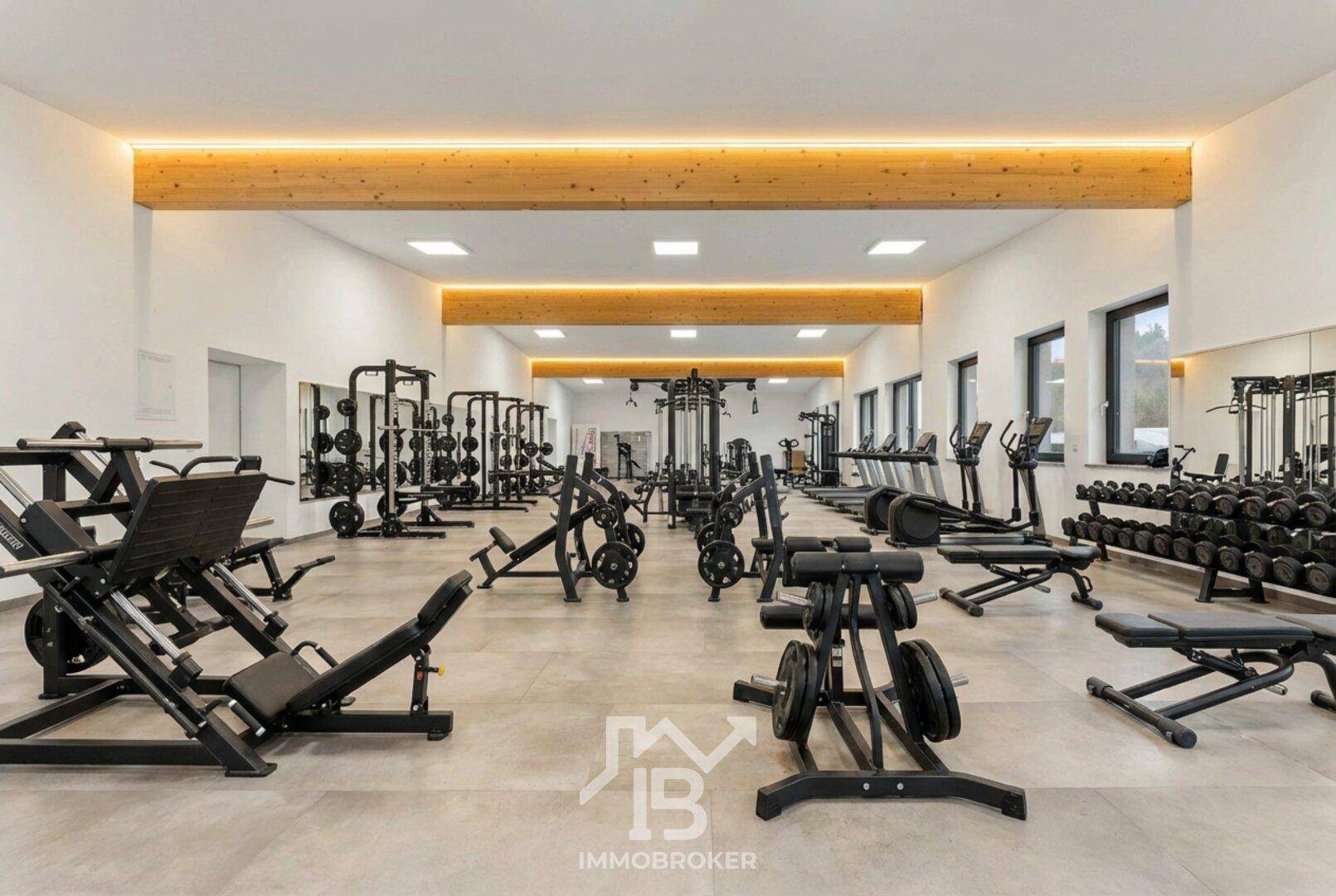 Moderner Fitnessraum mit einer Vielzahl von Kraft- und Cardiogeräten, großen Spiegeln und Holzbalken an der Decke.