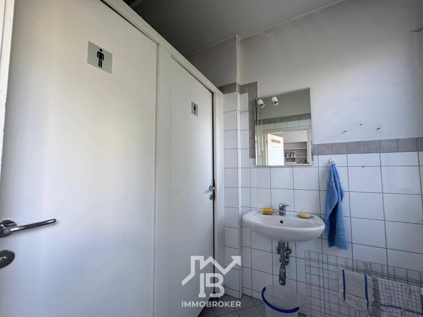 Sanitärbereich mit Waschbecken und zwei Toilettenkabinen, gefliesten Wänden und Spiegel.