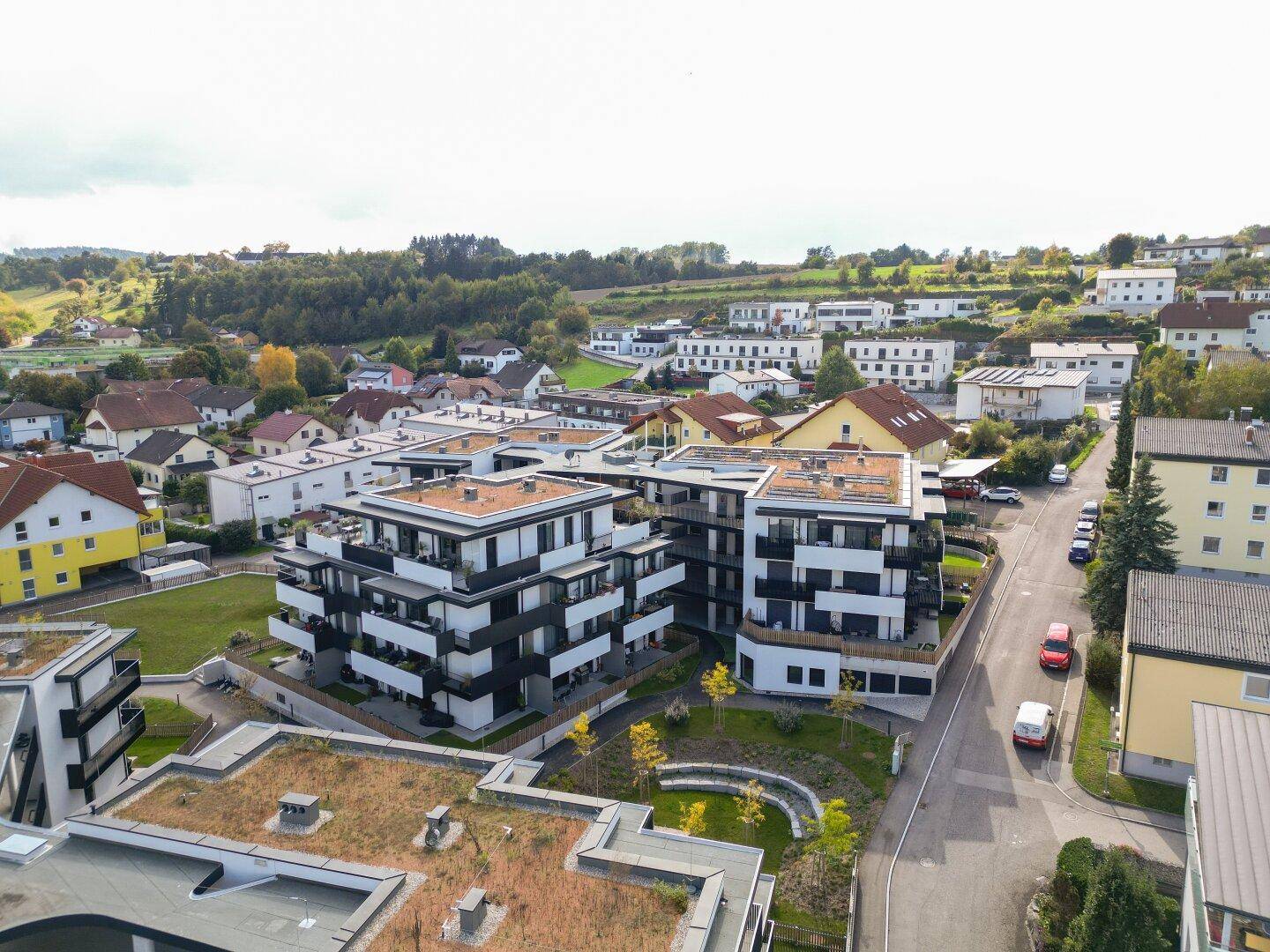 Luftaufnahme einer modernen Wohnanlage mit mehreren Gebäuden in einer grünen Hügellandschaft.
