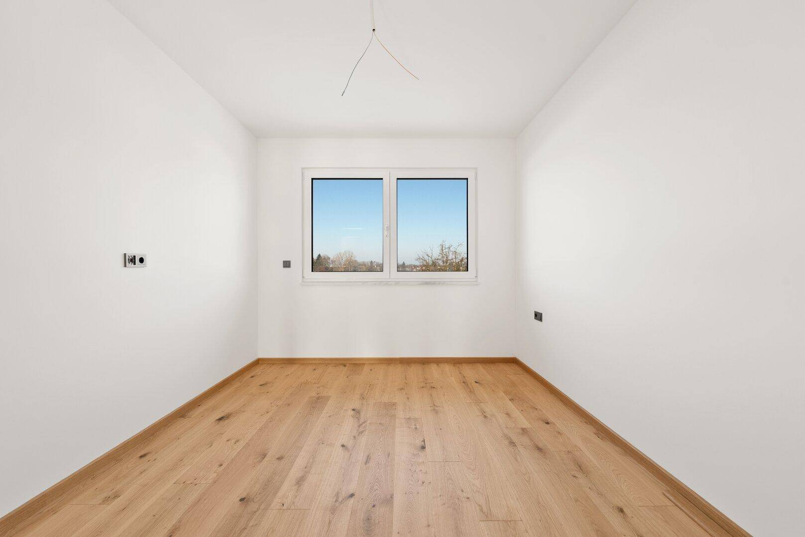 Helles Zimmer mit Parkettboden und einem großen Fenster, das viel Tageslicht hereinlässt.