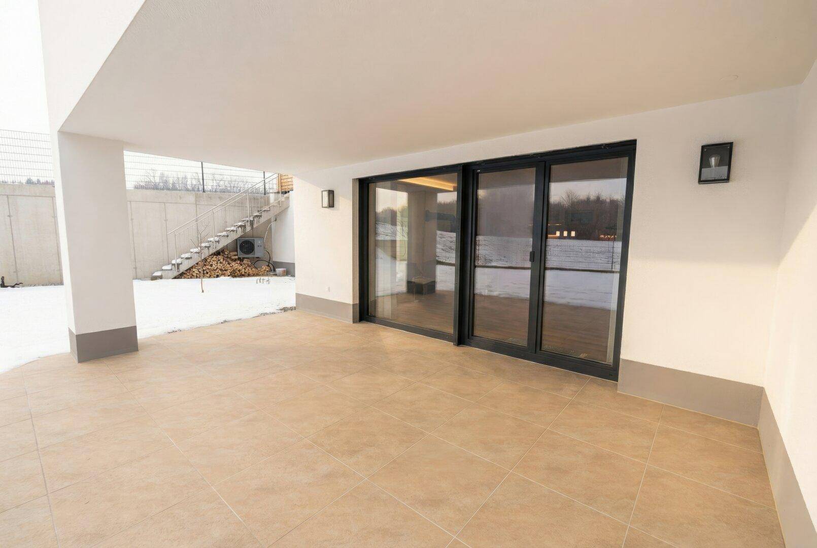 Überdachte Terrasse mit Fliesenboden und Zugang zu großen Glas-Schiebetüren, umgeben von Schnee.