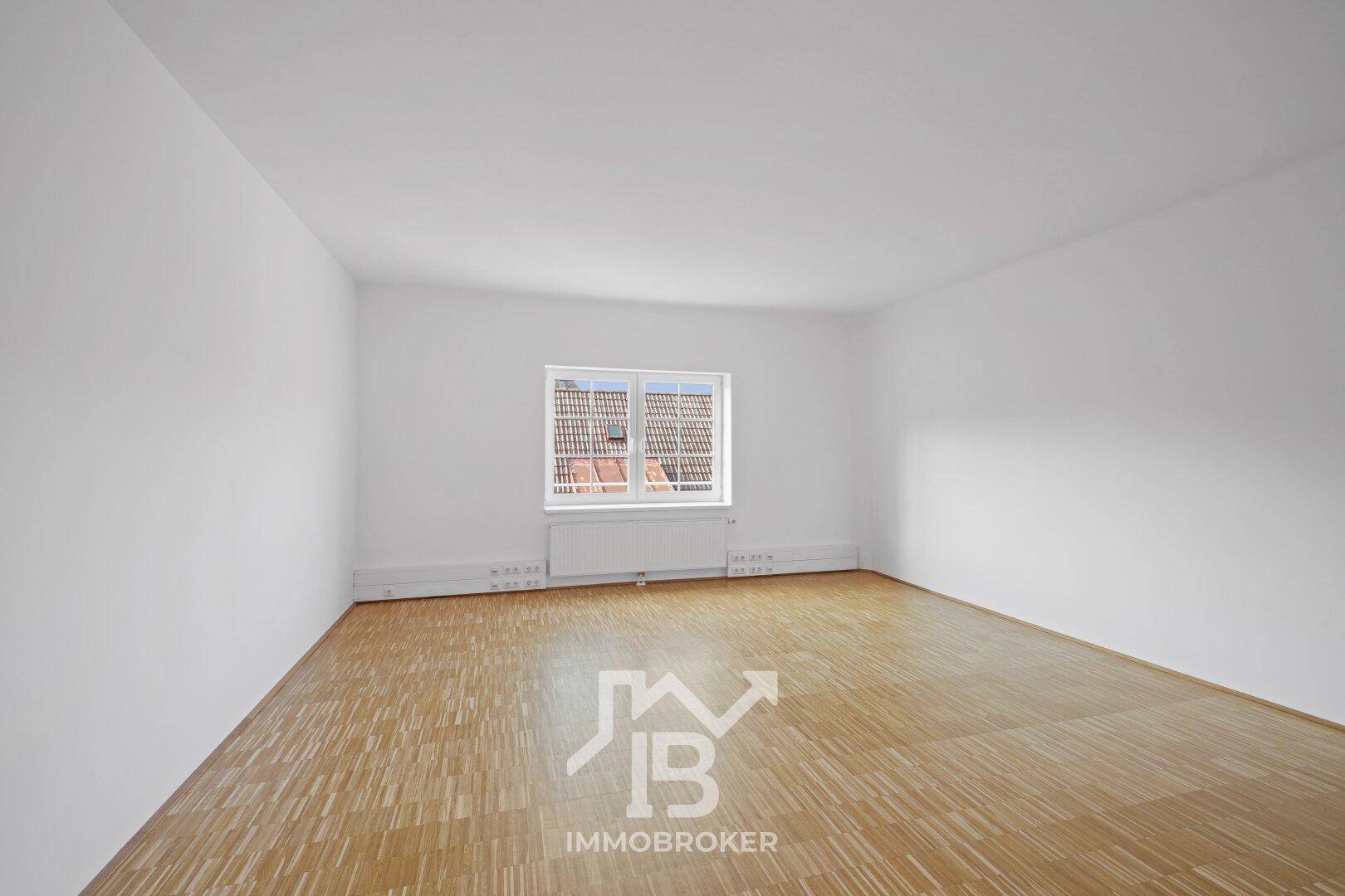 Heller Raum mit einem Fenster, Parkettboden und Heizkörpern, bietet einen Ausblick auf die umliegenden Dächer.