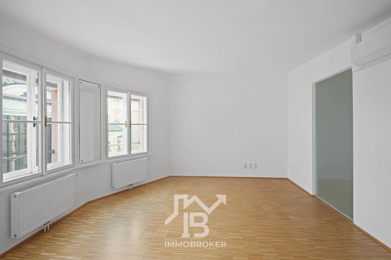 Heller Wohnraum mit großen Fenstern, Parkettboden und Heizkörpern, bietet einen Blick auf die umliegenden Gebäude.