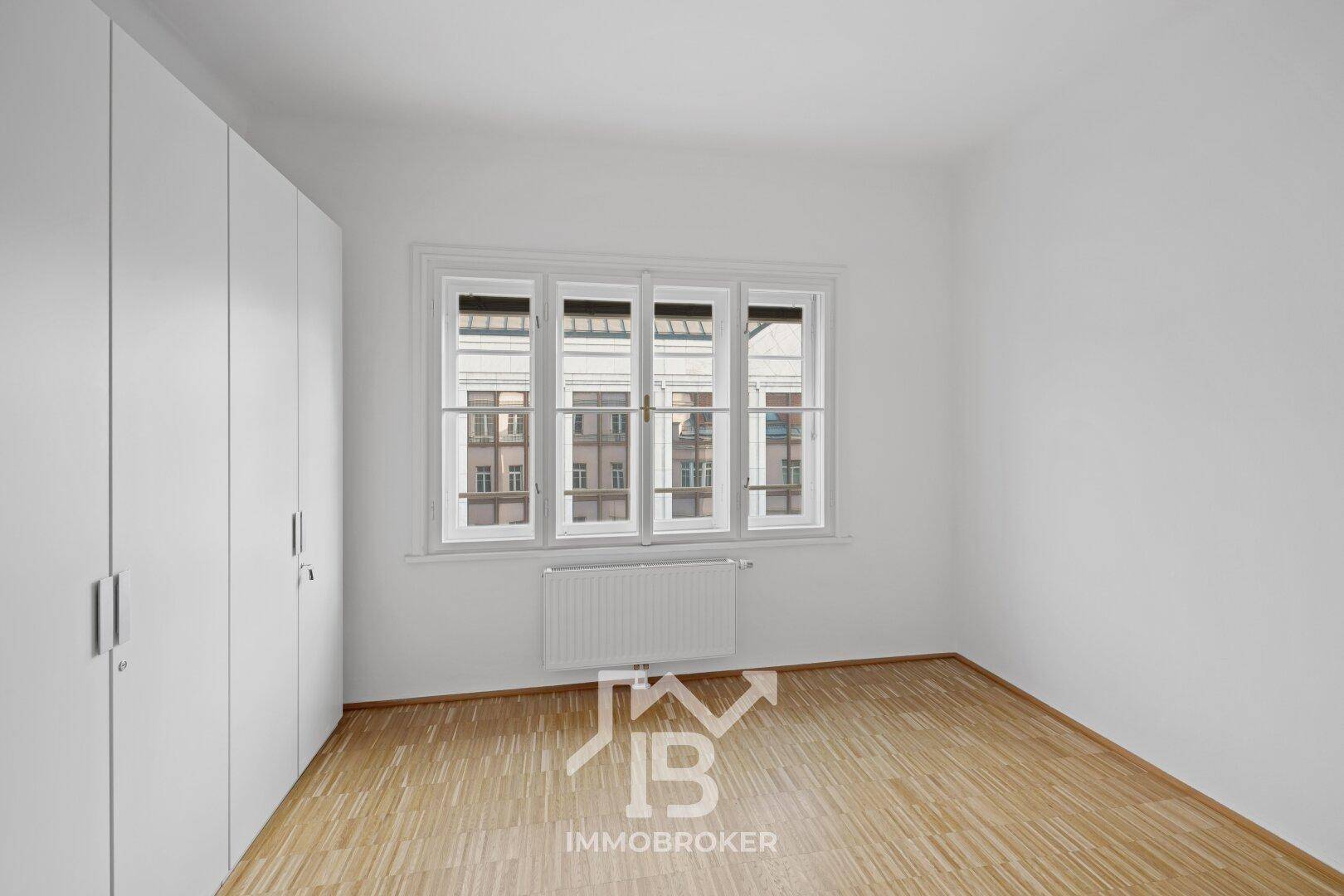 Helles Schlafzimmer mit großem Fenster, Parkettboden und einem praktischen weißen Einbauschrank.