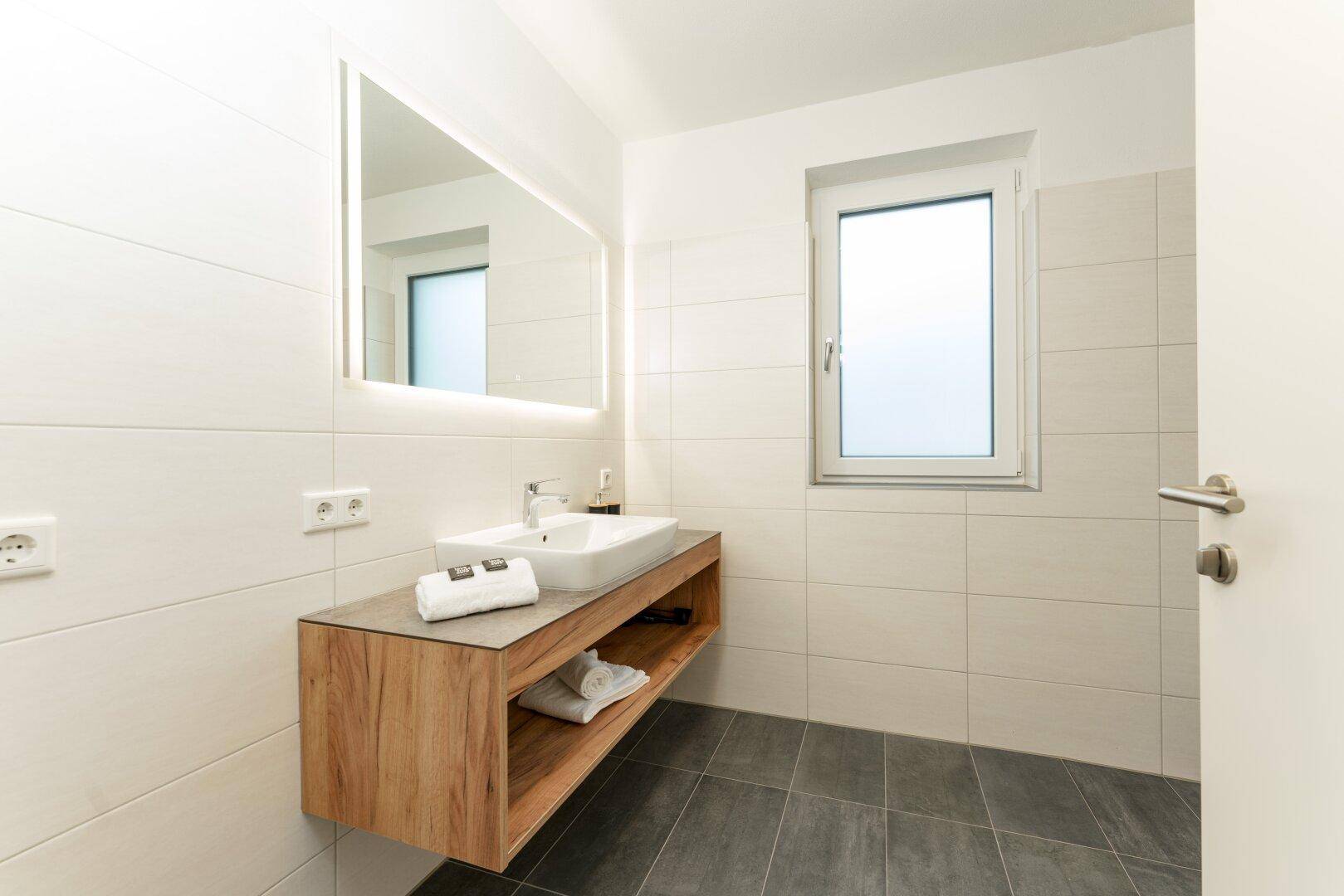Modernes Badezimmer mit Waschtisch, beleuchtetem Spiegel und Fenster für Tageslicht.