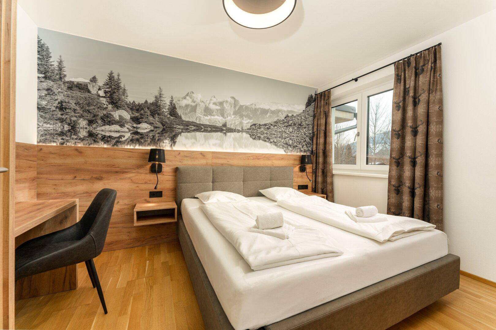 Helles Schlafzimmer mit Doppelbett, Holzboden und großem Wandbild einer Berglandschaft.