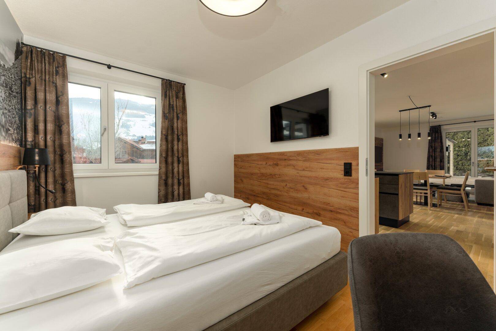 Schlafzimmer mit Doppelbett, Fernseher und Zugang zum offenen Wohn-Essbereich.
