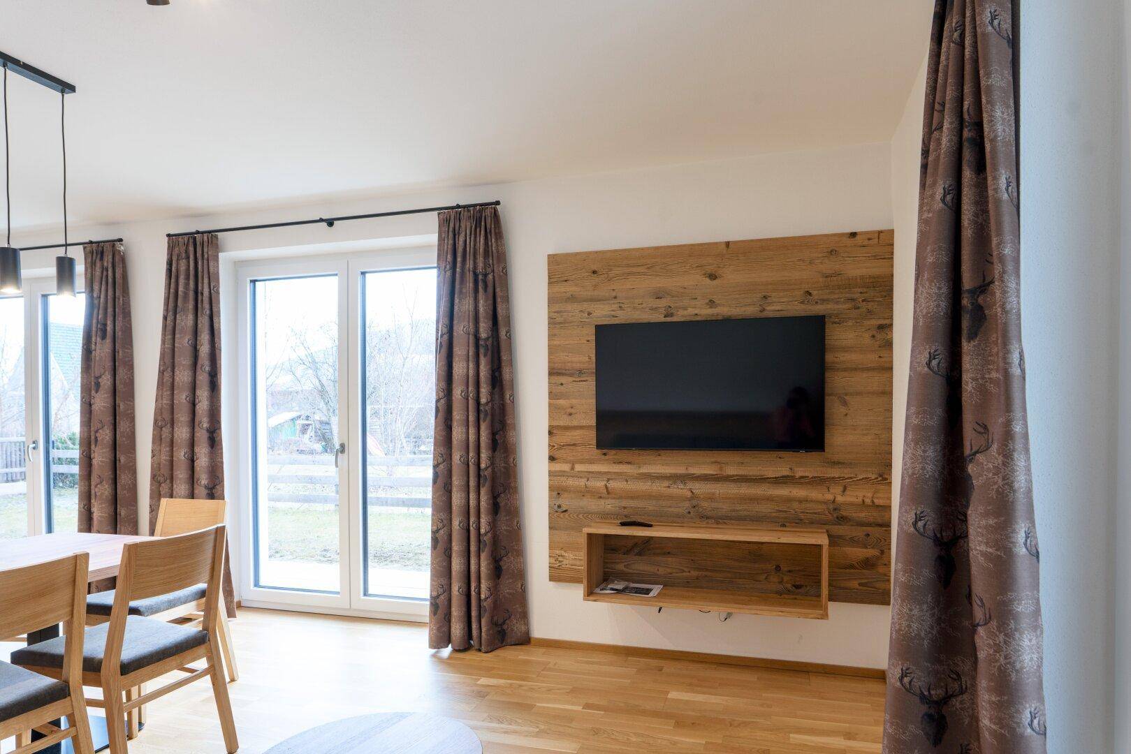 Wohnbereich mit Holzwand, Flachbildfernseher und großen Fenstern mit Blick ins Freie.