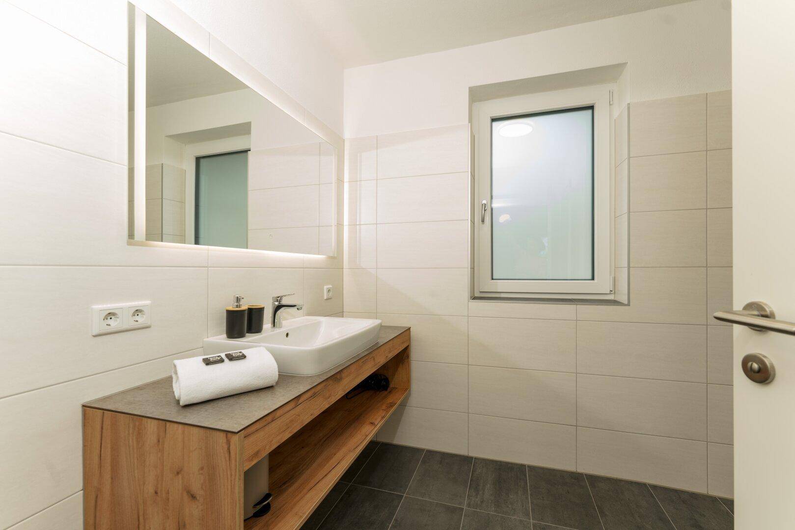 Modernes Badezimmer mit Einzelwaschbecken, großem Spiegel und Fenster für natürliches Licht.