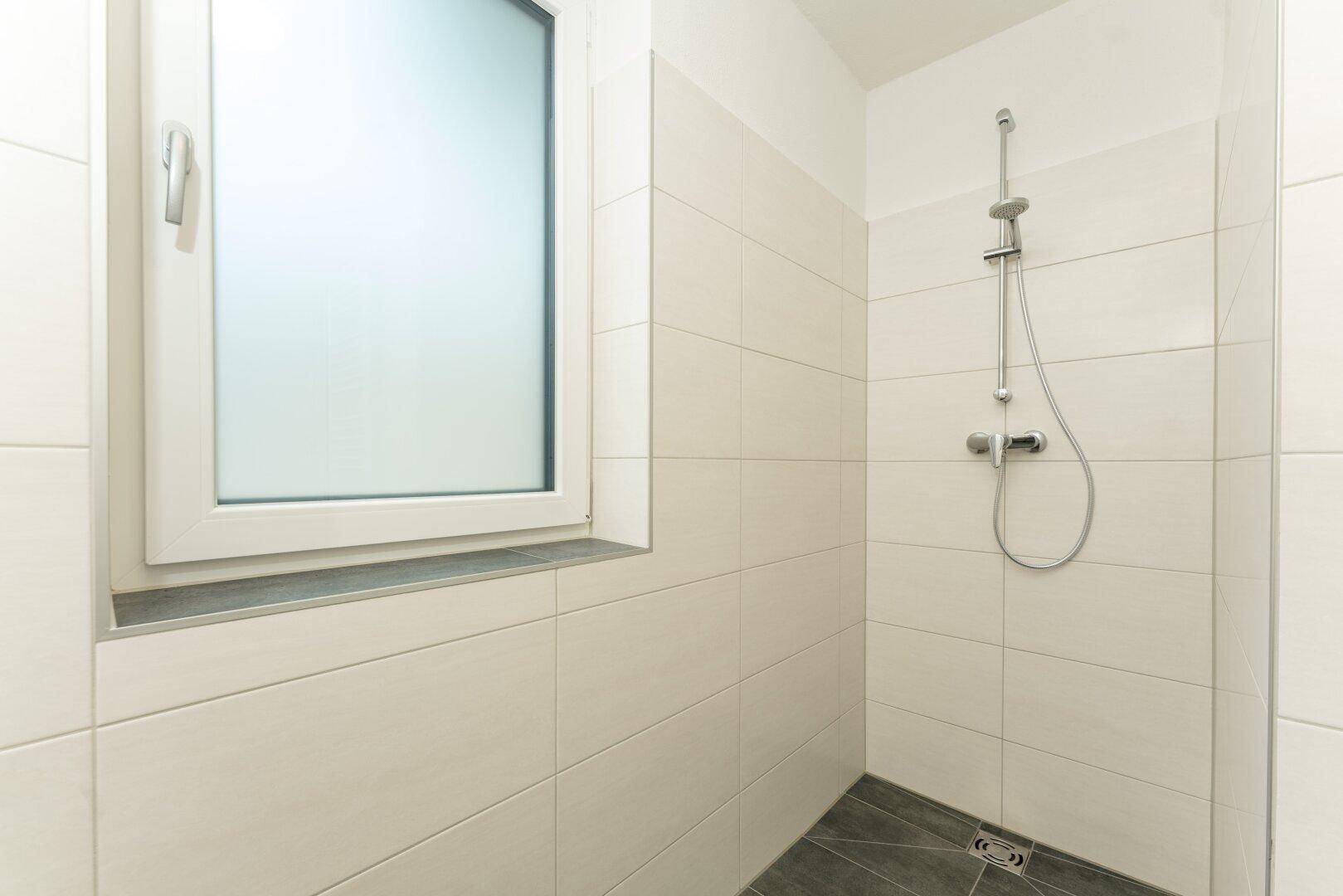Duschbereich im Badezimmer mit weißen Fliesen und einem Fenster für gute Belüftung.