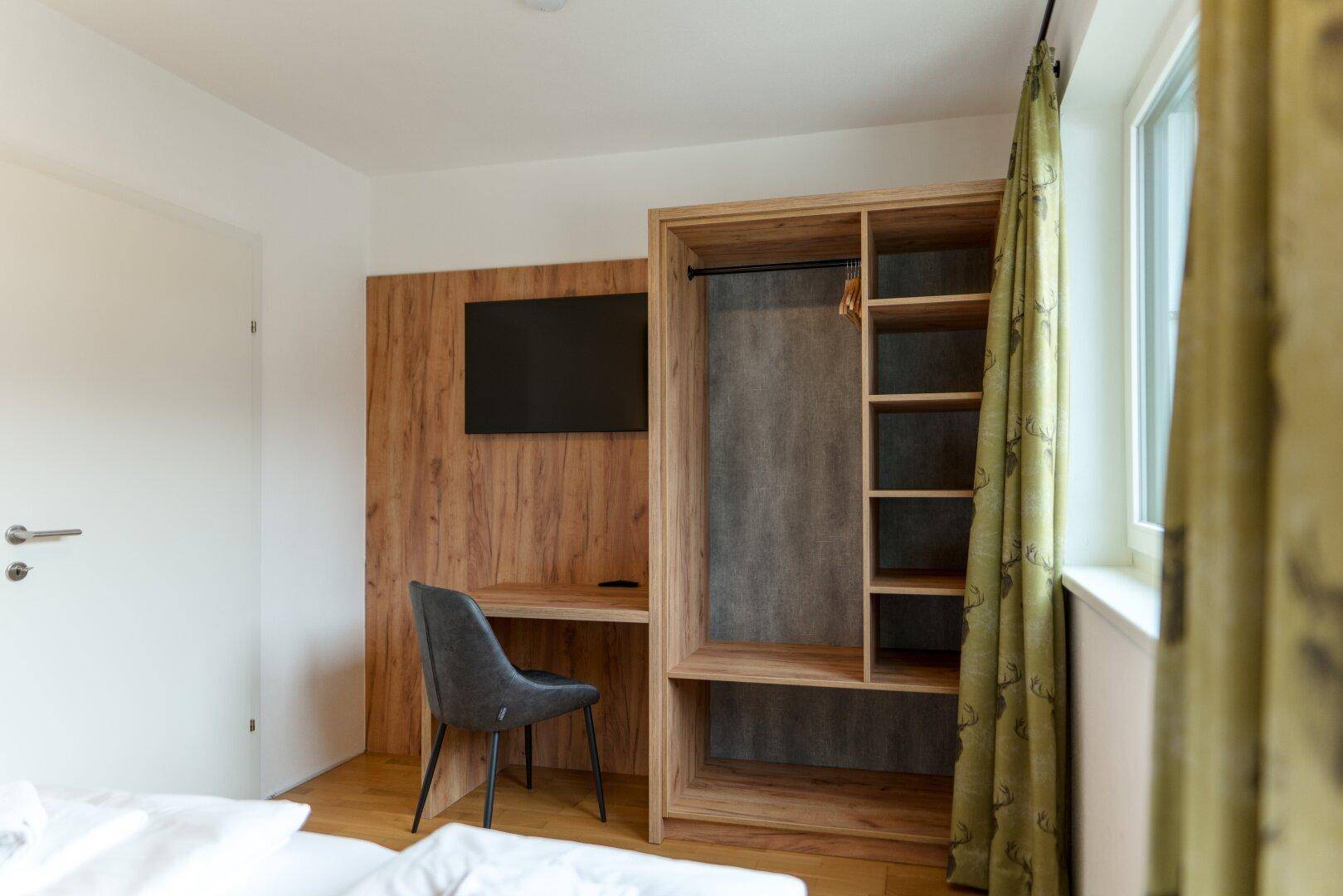 Arbeitsbereich im Schlafzimmer mit Schreibtisch, Fernseher und offenem Kleiderschrank in Holzoptik.