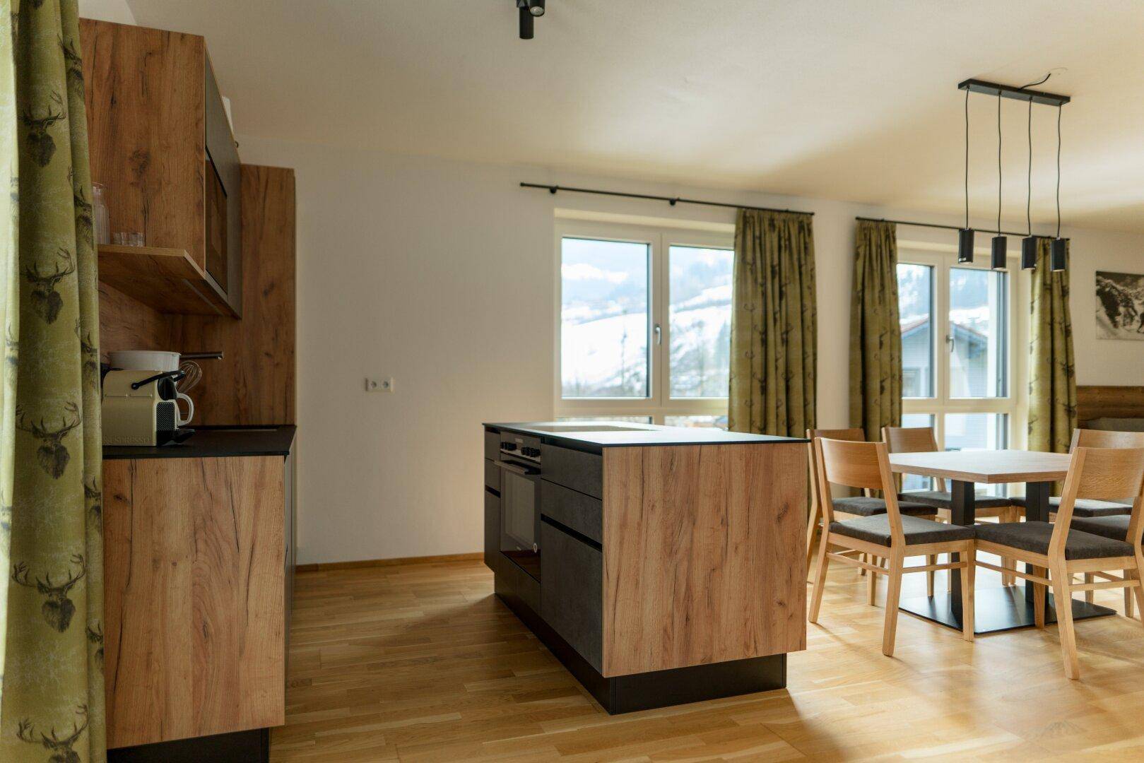 Küchenbereich mit Kochinsel und Blick auf den Esstisch sowie die großen Fenster mit Bergblick.