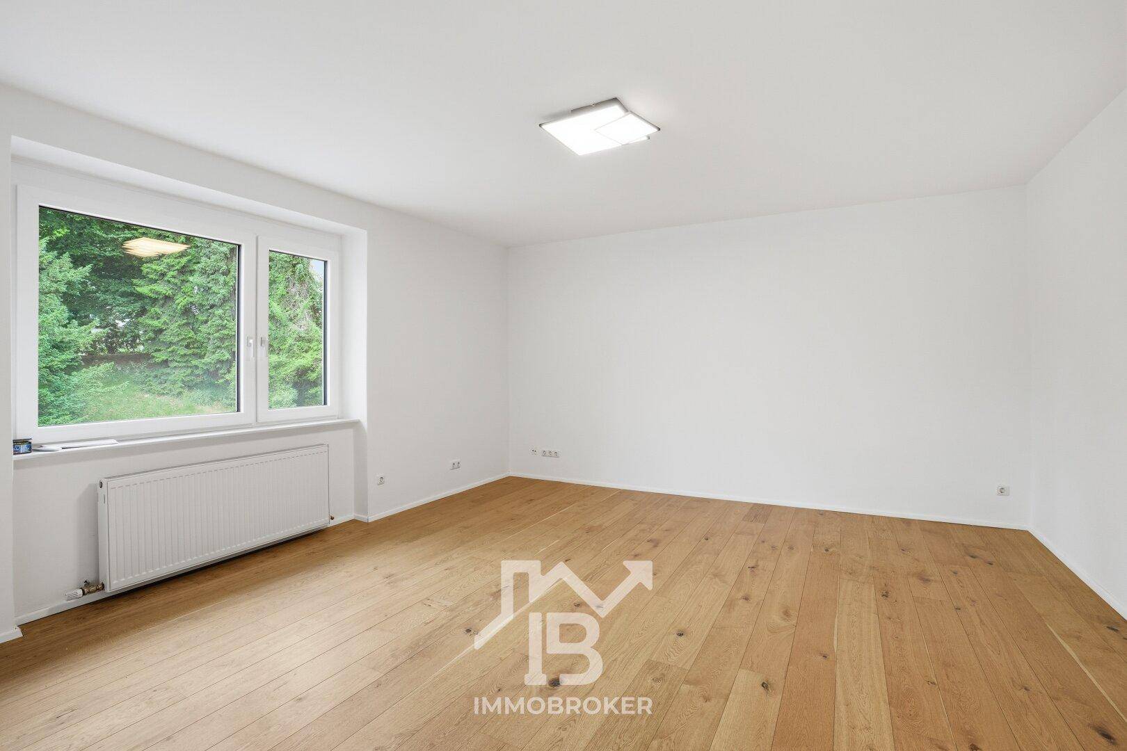 Geräumiges Zimmer mit hellem Holzboden und Fenster mit Blick auf Bäume.
