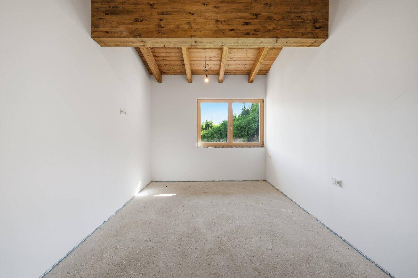 Unfertiger Raum mit Holzbalkendecke, weißen Wänden und einem Fenster mit Blick ins Grüne.