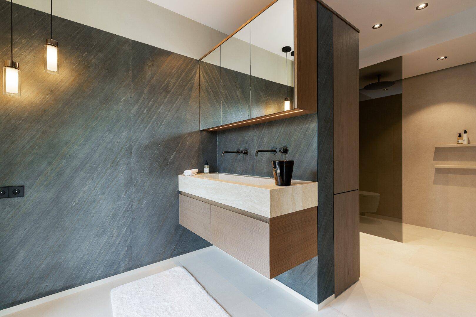 Elegantes Badezimmer mit dunklen Steinfliesen, schwebendem Waschtisch und großem Spiegelschrank.