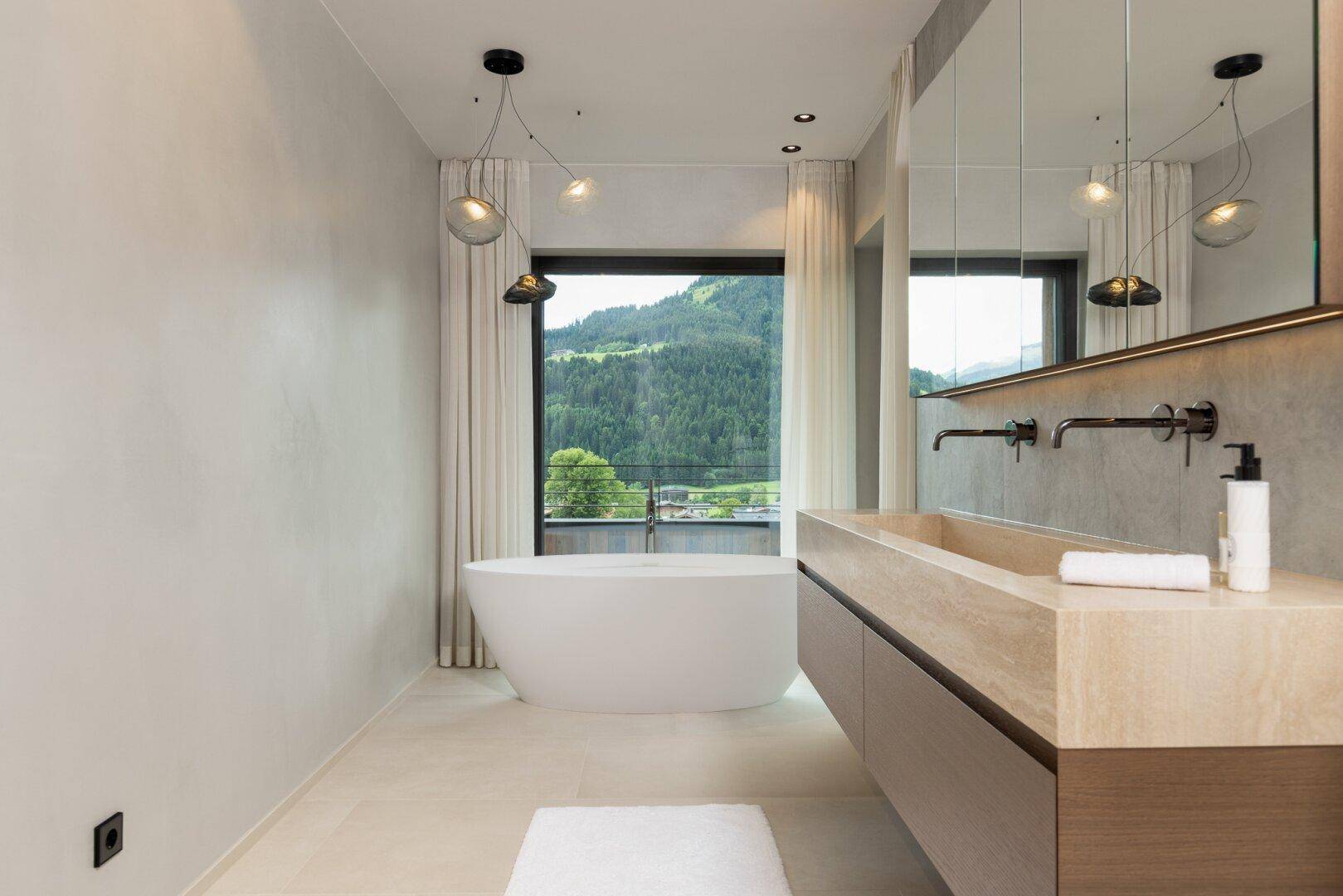 Luxuriöses Badezimmer mit freistehender Badewanne, Doppelwaschtisch und Panoramafenstern mit Bergblick.