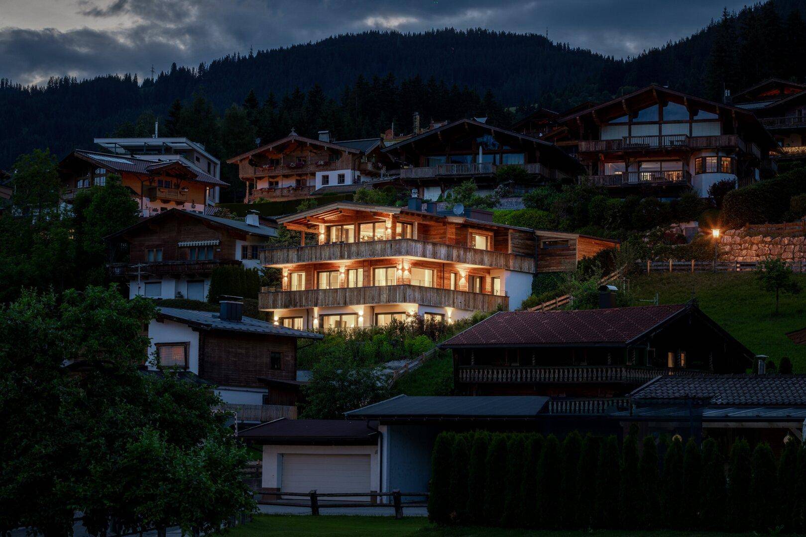 Beleuchtetes Chalet in den Bergen bei Nacht, umgeben von weiteren Häusern und viel Grün.