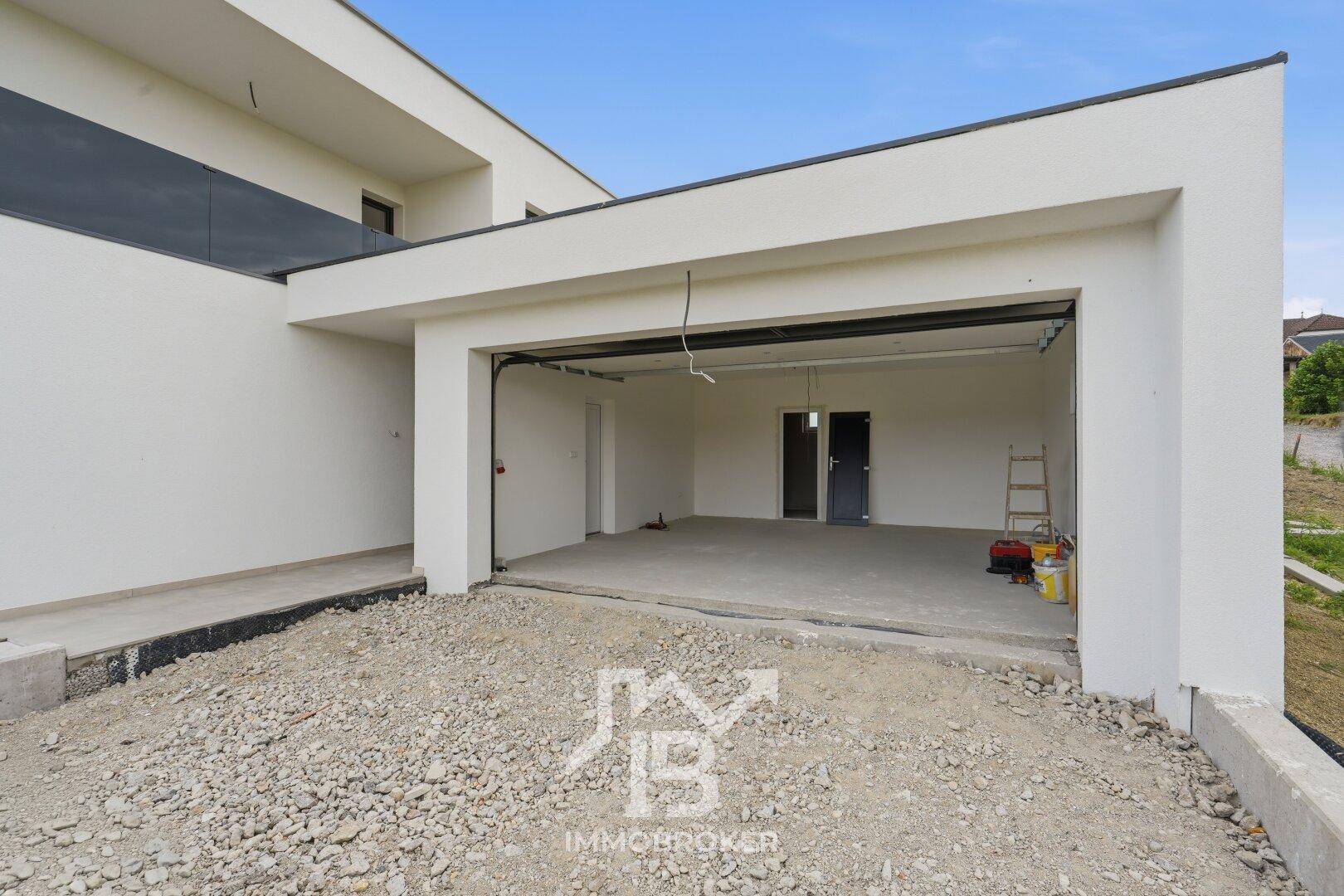 Offene Garage eines modernen Hauses mit Blick auf den leeren Innenraum und Schotterzufahrt.