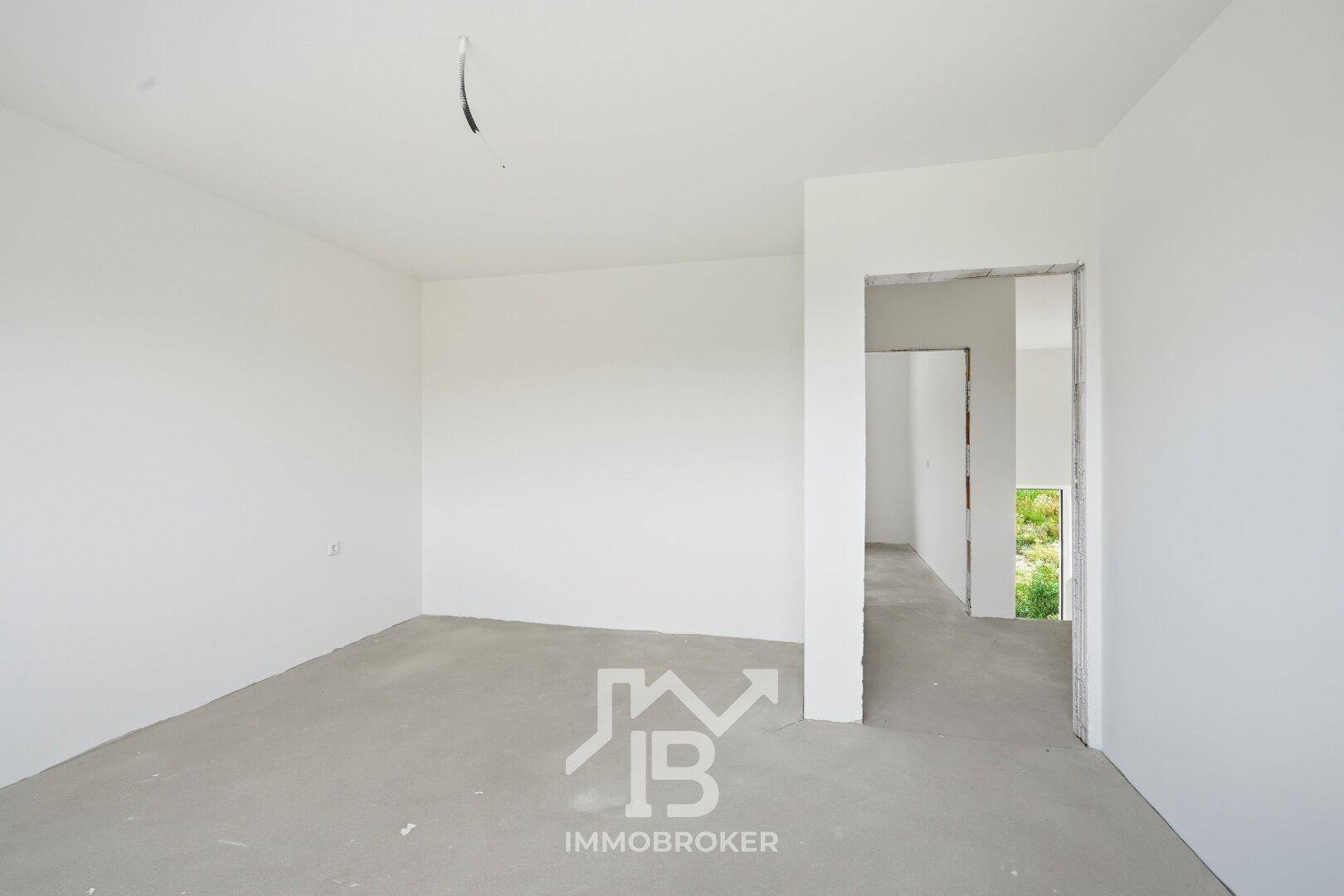 Unfertiger Raum mit Betonboden und offenem Durchgang zu einem weiteren Rohbaubereich.