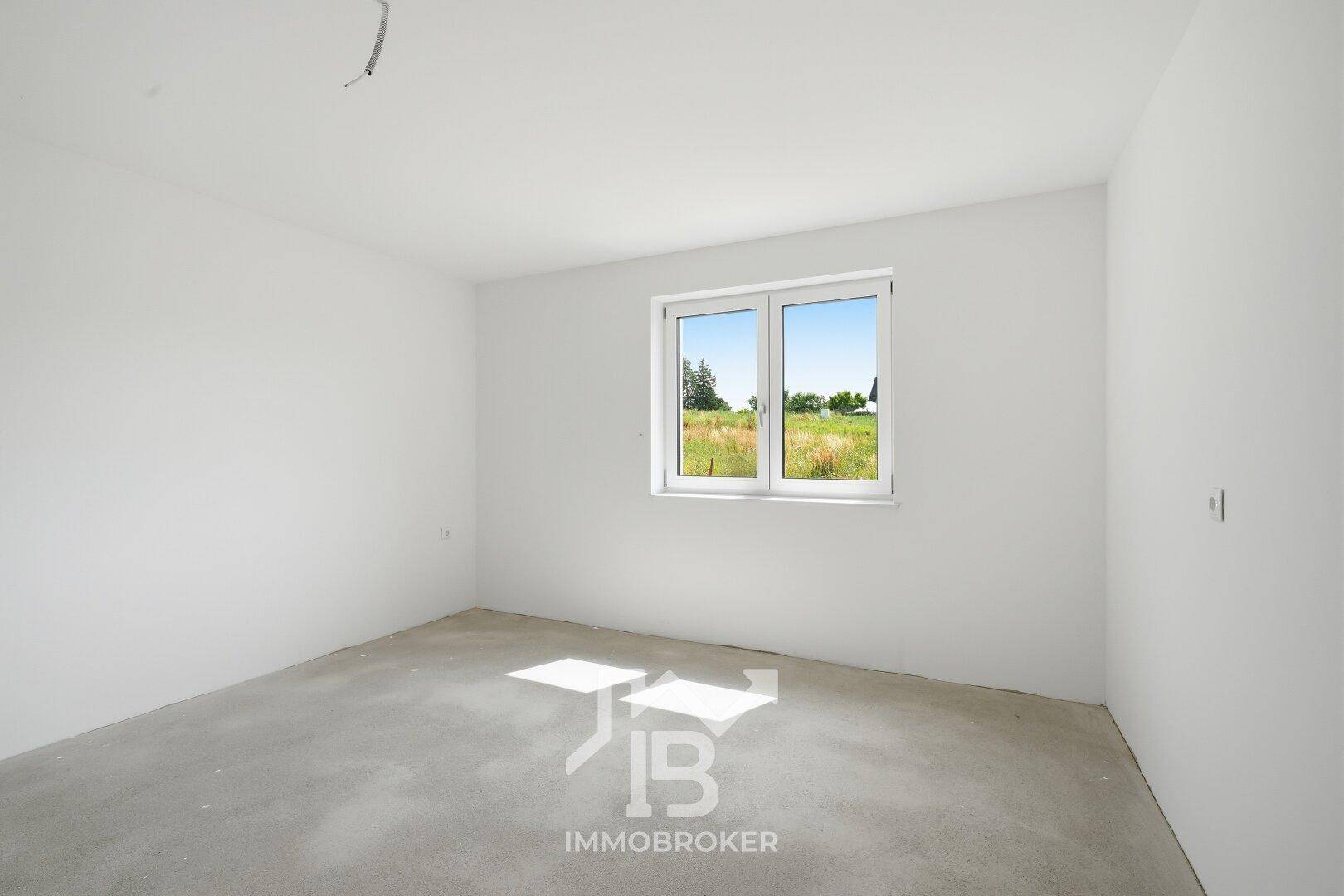 Heller Raum im Rohbau mit Betonboden und Fenster, das einen Blick auf die Felder bietet.