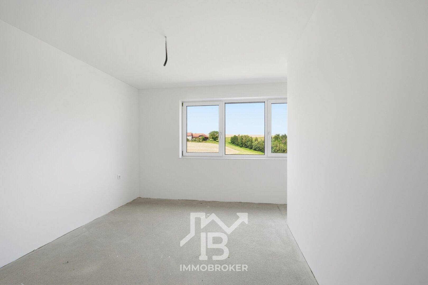 Geräumiger Raum im Rohbau mit Betonboden und großem Fenster, das viel Tageslicht hereinlässt.