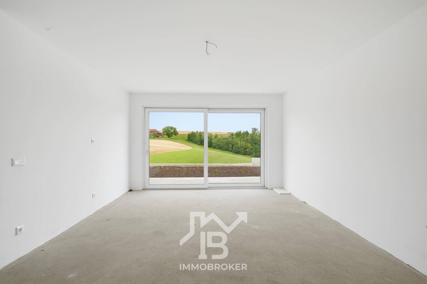 Unfertiger Wohnraum mit Betonboden und großen Fenstern, die einen weiten Blick ins Grüne bieten.