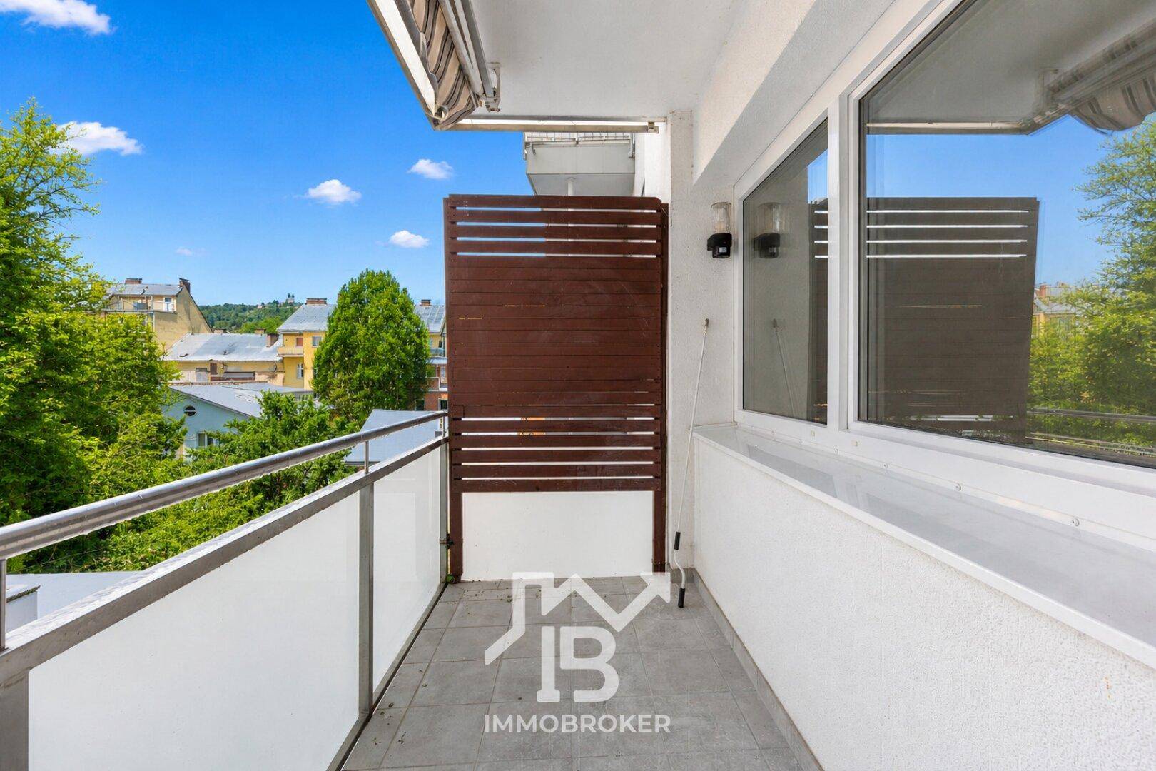 Heller Balkon mit Fliesenboden, Glasbrüstung und Markise, bietet Blick ins Grüne.