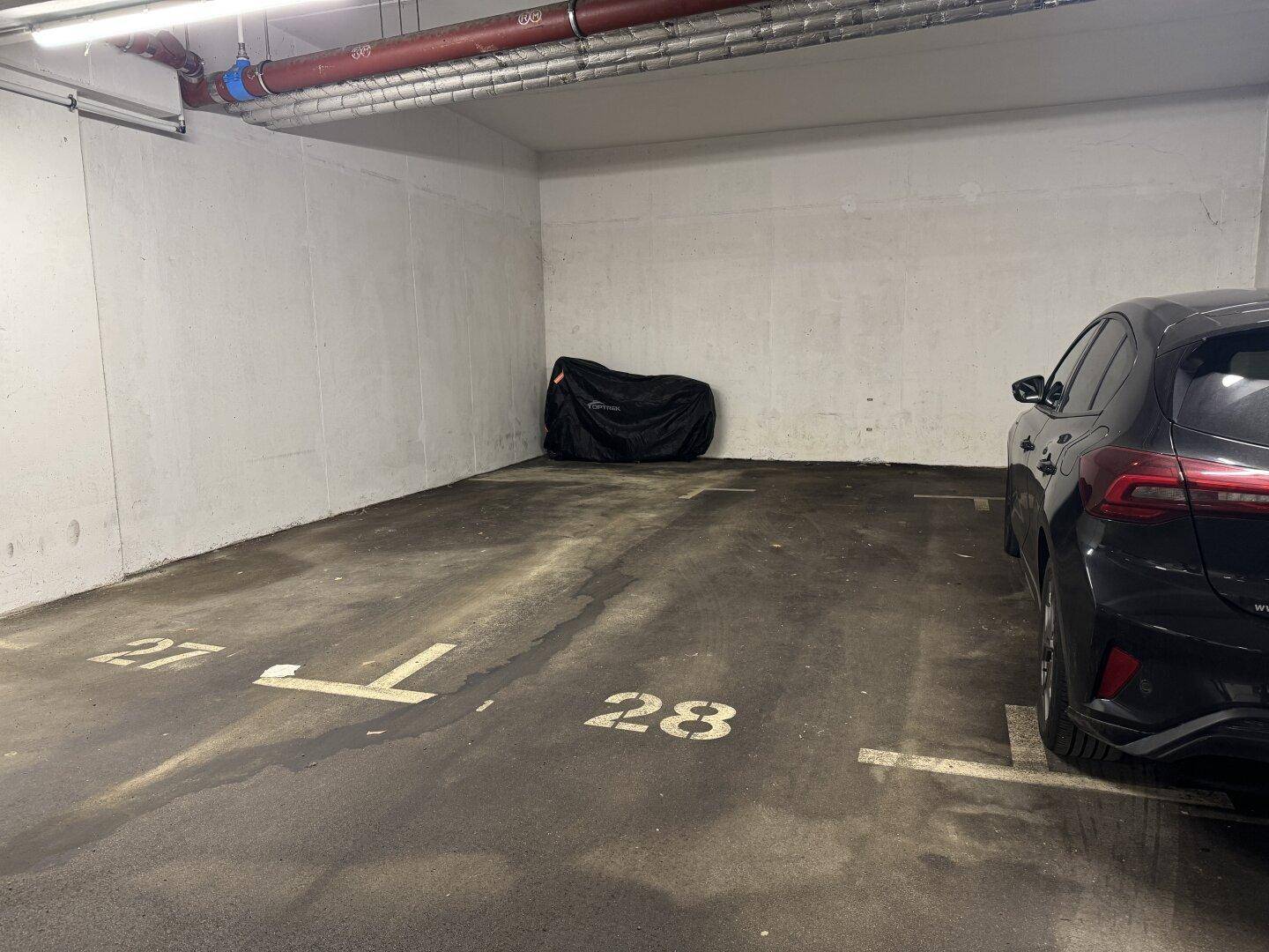 Tiefgarage mit markierten Parkplätzen, einem geparkten Auto und sichtbaren Versorgungsleitungen an der Decke.