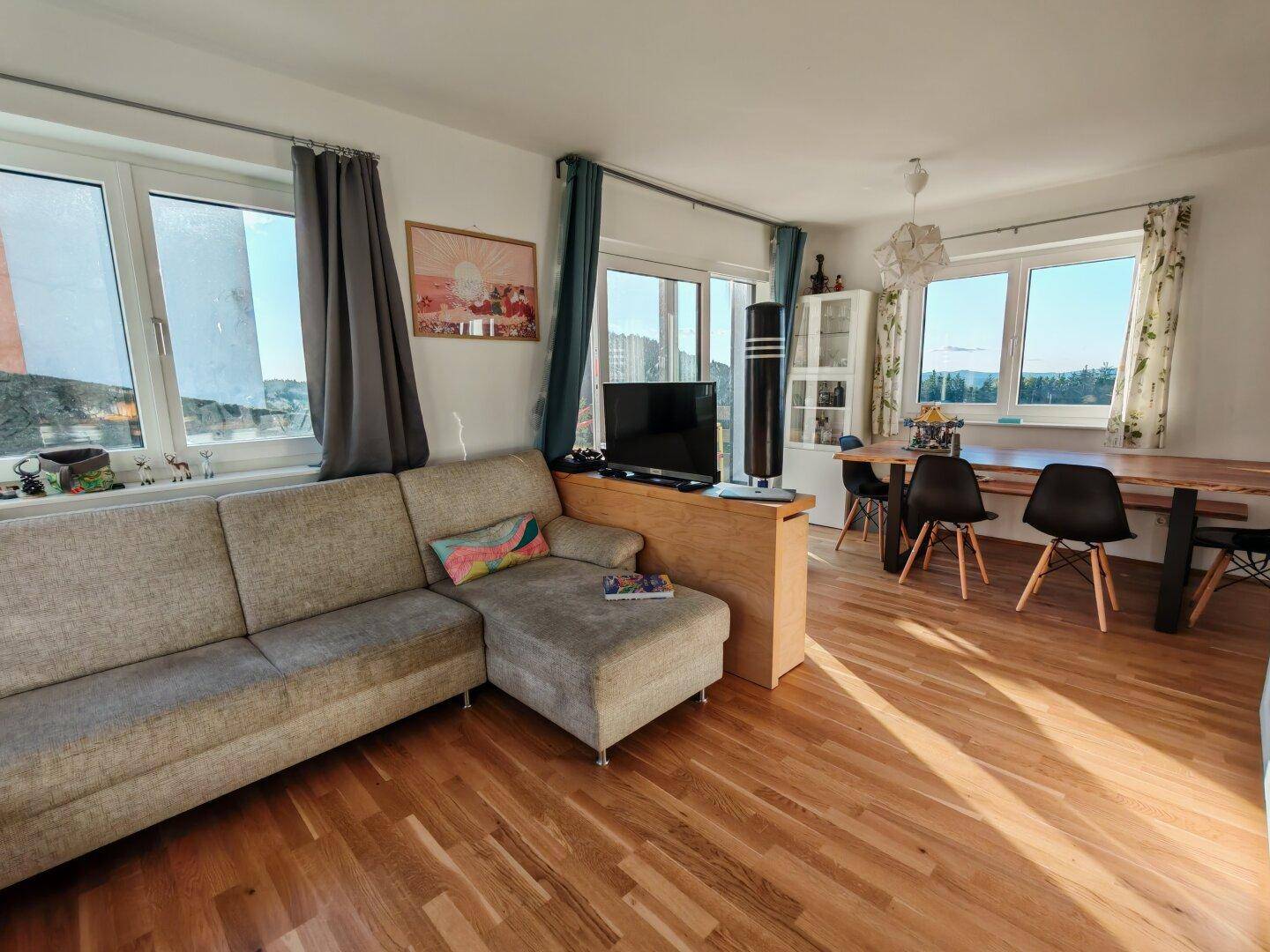 Gemütlicher Wohn-Essbereich mit Sofa, Holzboden und großzügigen Fenstern mit Ausblick ins Grüne.
