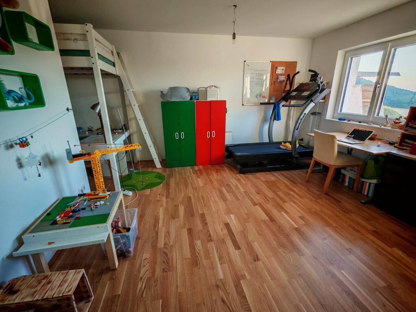 Kinderzimmer mit Hochbett, Schreibtisch und Holzboden, hell durch große Fenster mit Ausblick.