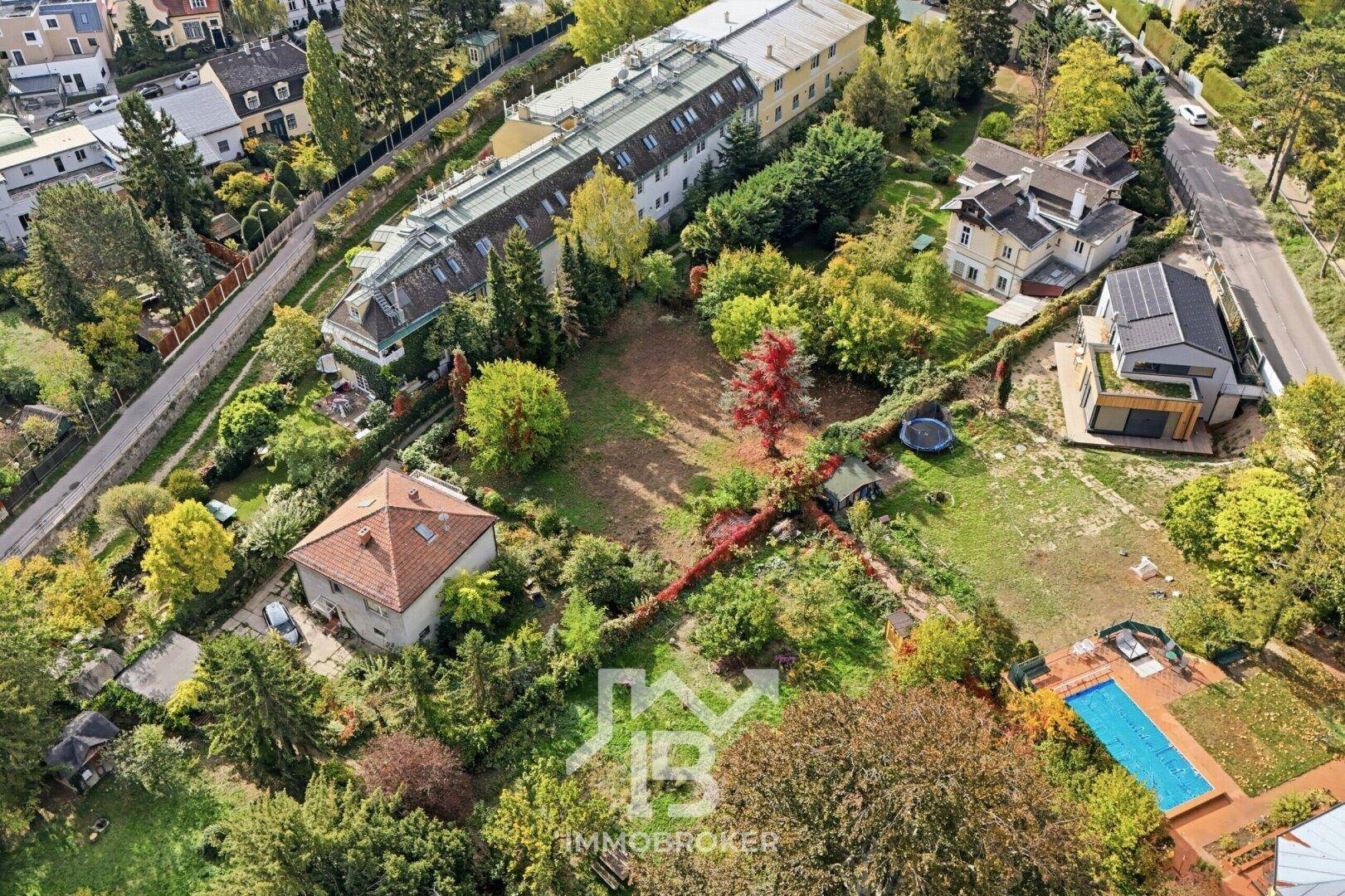Weitläufige Luftaufnahme eines unbebauten Grundstücks neben einer modernen Villa und weiteren Häusern.