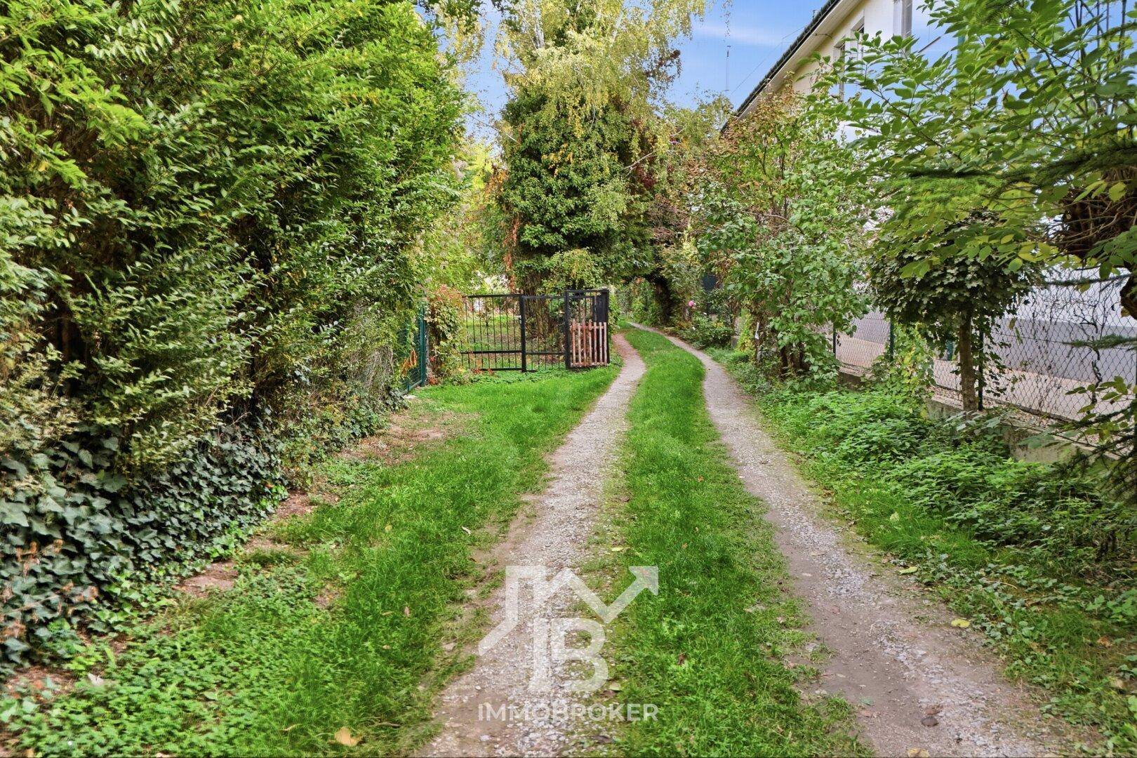 Schotterweg führt durch dichte Hecken und Bäume zu einem Gartentor, umgeben von Natur.