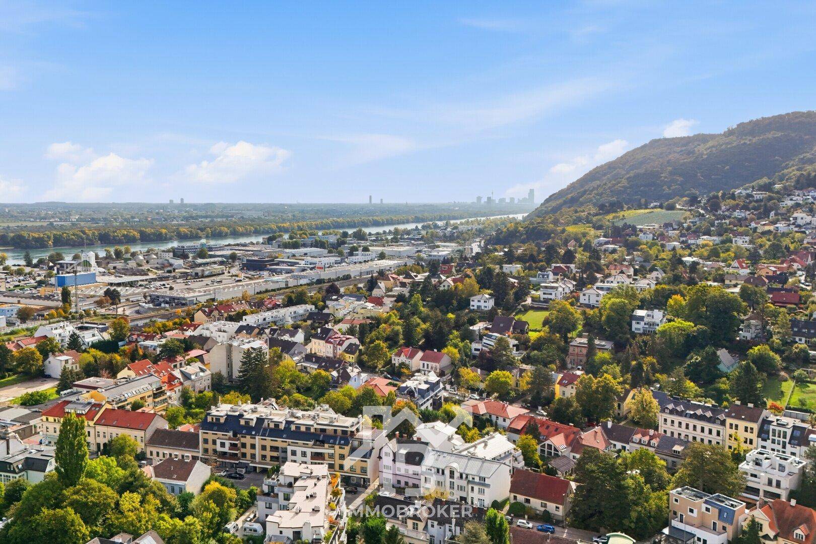 Weitläufiger Blick über eine Stadt mit Fluss, grünen Hügeln und urbaner Skyline am Horizont.