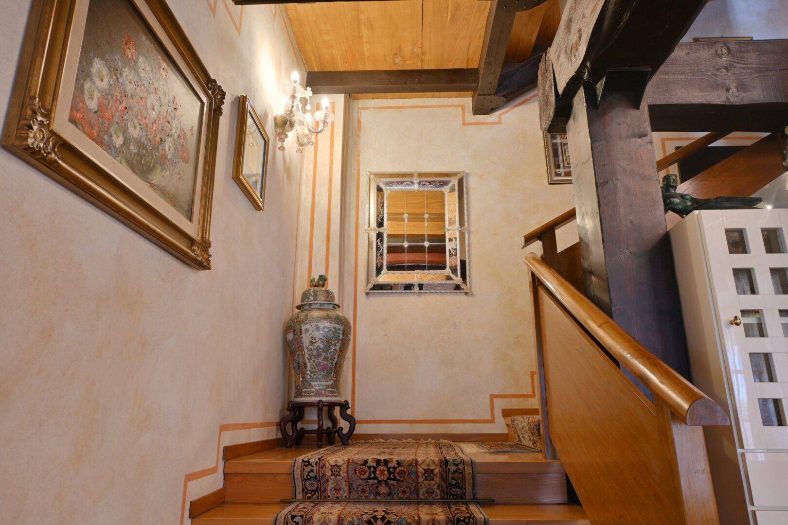 Helles Treppenhaus mit Holzbalkendecke, gemustertem Teppich und klassischer Wanddekoration.