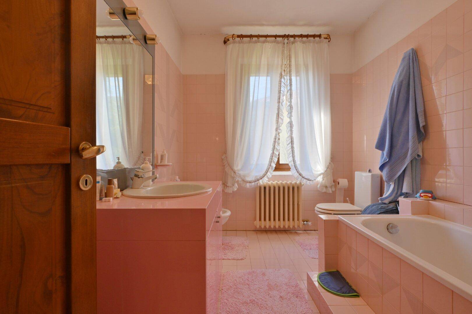 Badezimmer mit rosa Fliesen, Badewanne und Fenster mit Heizkörper.