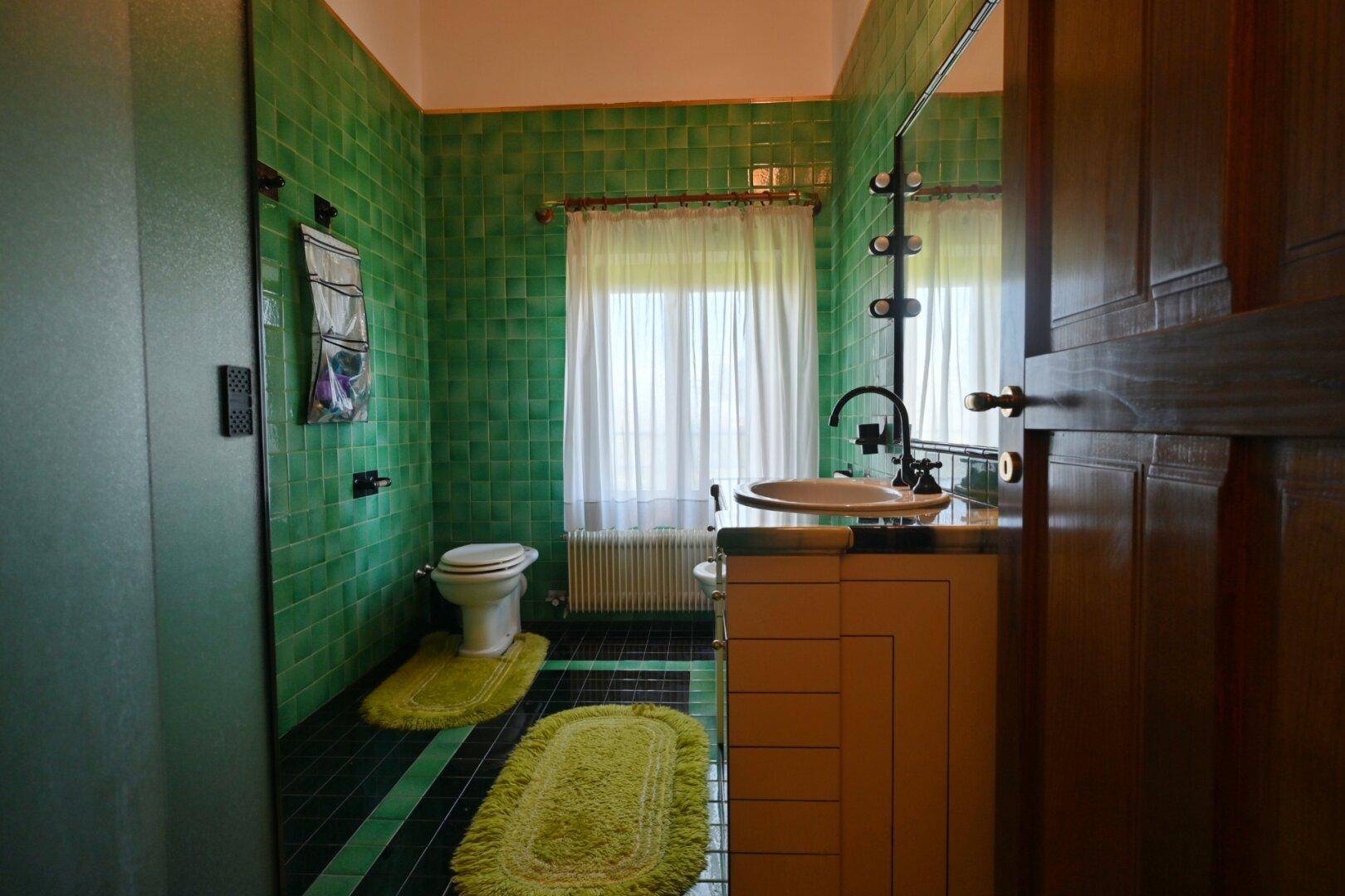 Badezimmer mit grünen Fliesen, Duschkabine und Fenster mit Heizkörper.