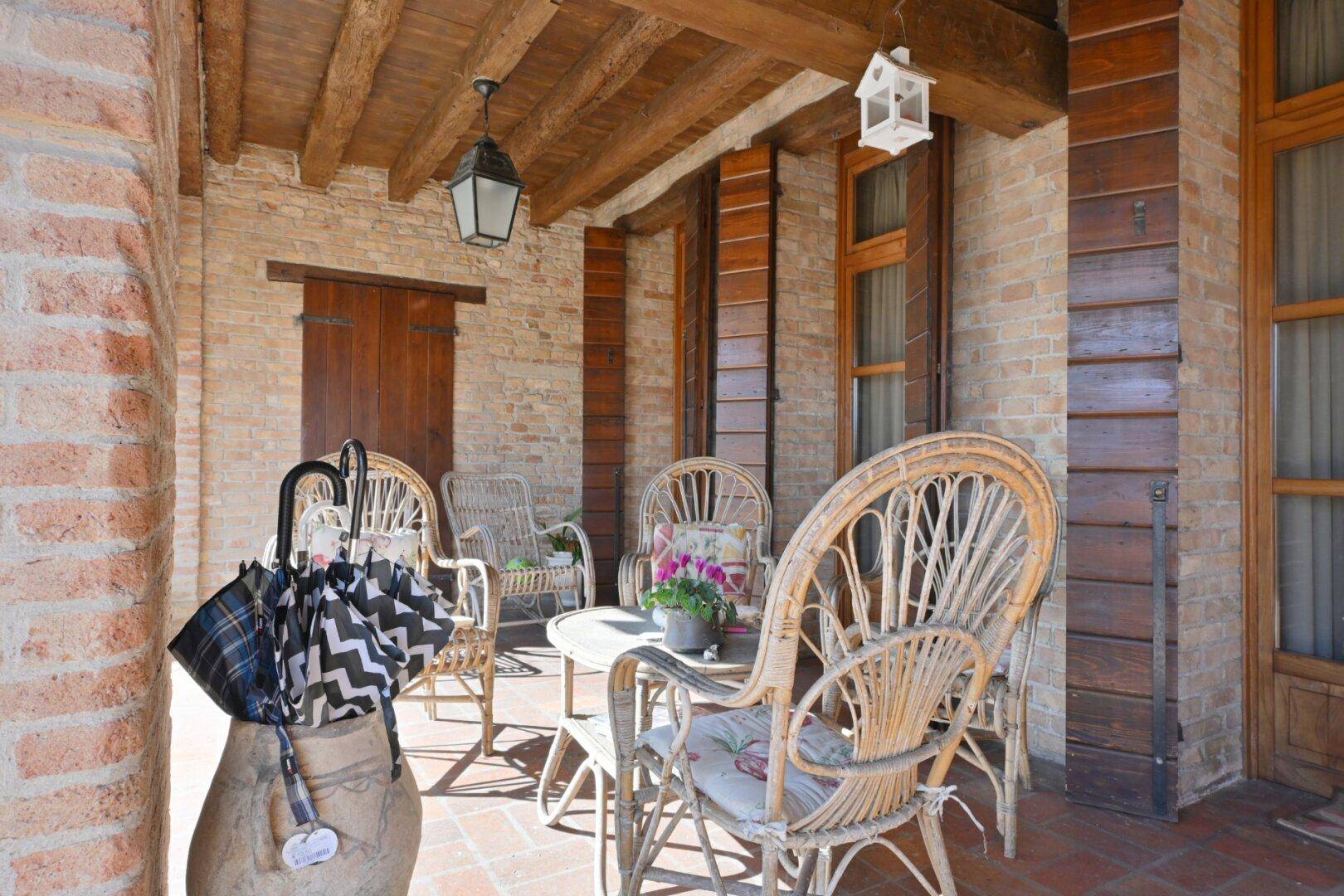 Überdachte Terrasse mit Korbmöbeln, Ziegelwänden und Holzbalkendecke.