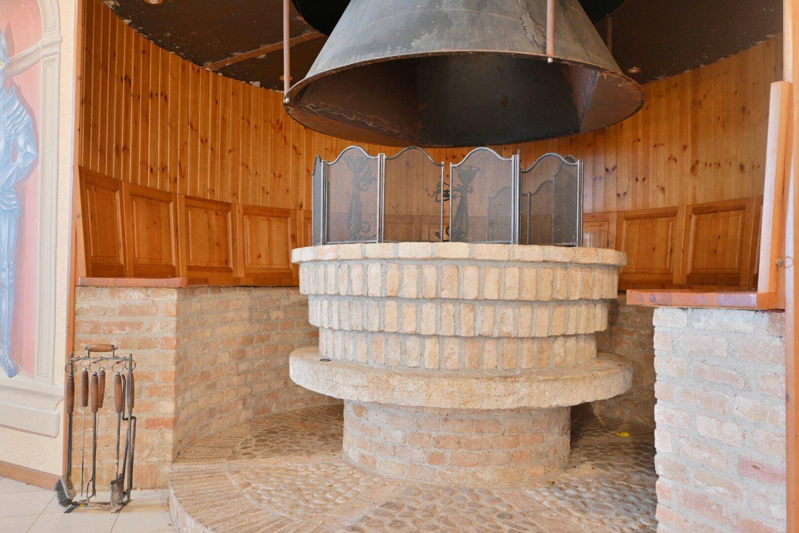 Großer, runder Kamin aus Ziegeln mit Metallhaube und Holzverkleidung.