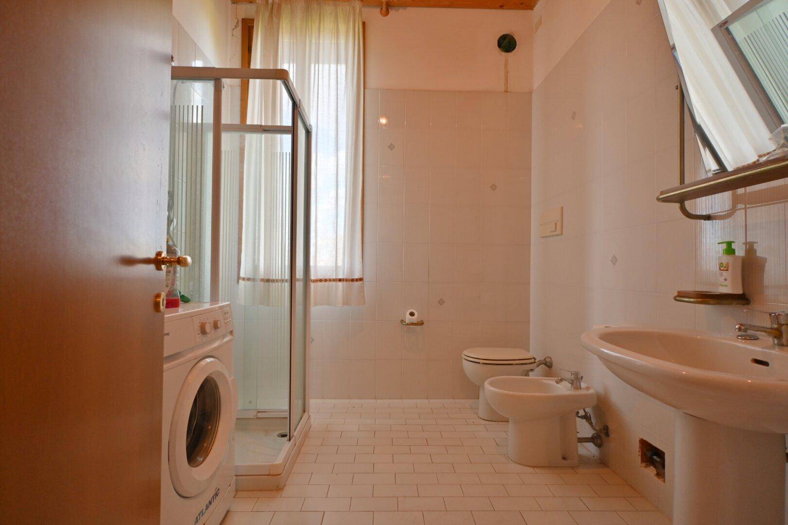 Helles Badezimmer mit Dusche, Bidet, Waschmaschine und weiß gefliesten Wänden.
