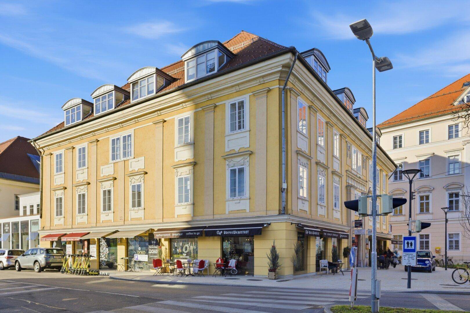 Eckgebäude mit gelber Fassade und Geschäften im Erdgeschoss, umgeben von städtischer Infrastruktur und Fußgängerzone.