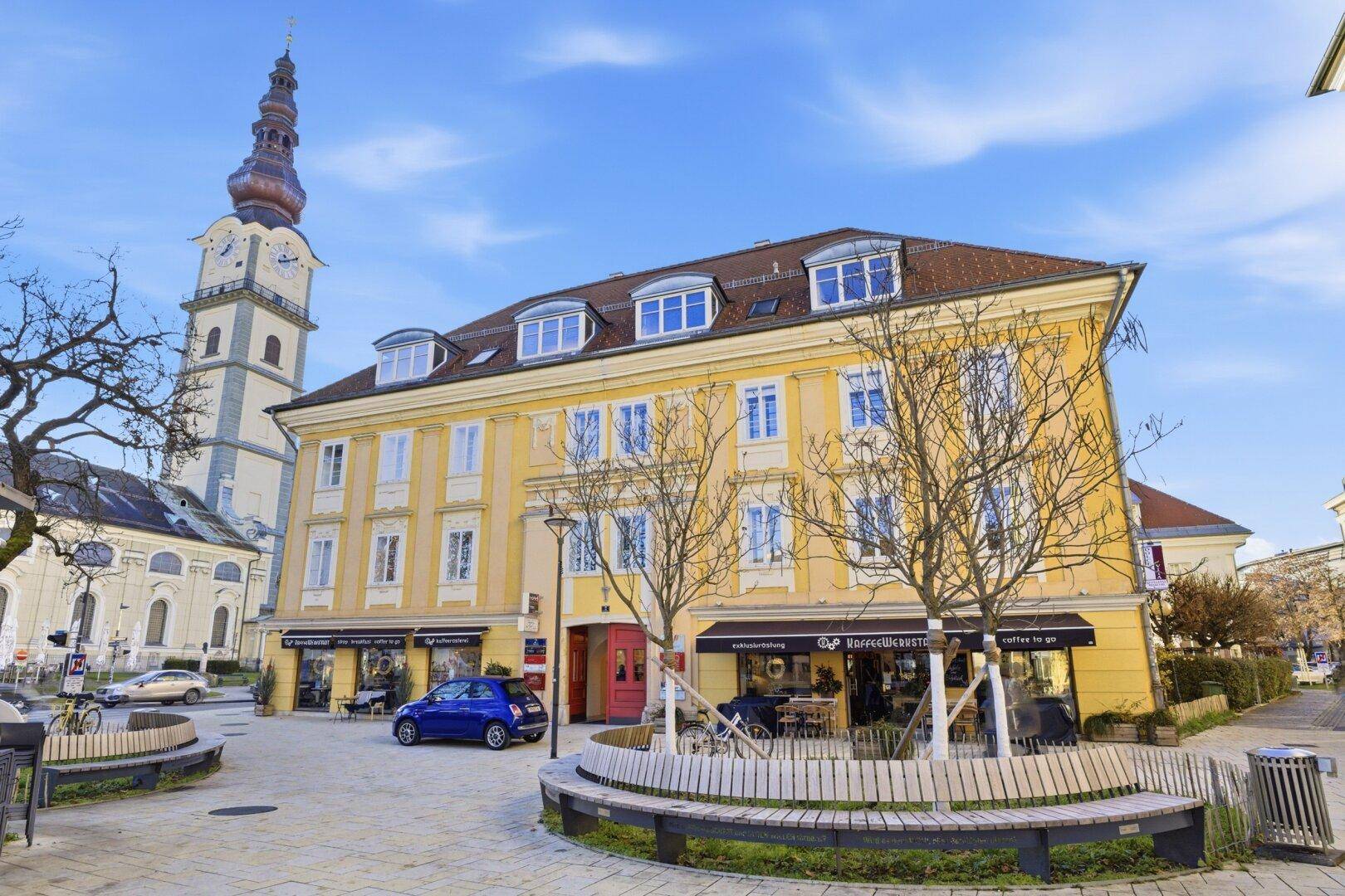 Historisches Gebäude mit gelber Fassade an einem Stadtplatz mit Kirche, Bäumen und Sitzgelegenheiten unter blauem Himmel.