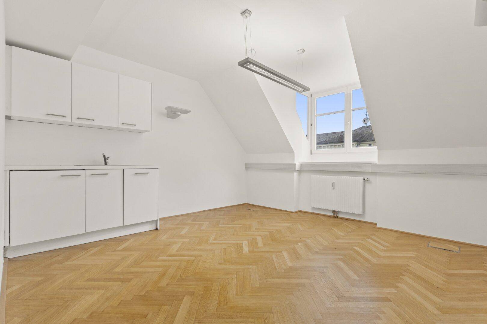 Heller Büroraum im Dachgeschoss mit kleiner Küchenzeile, Dachschrägen und Fischgrätparkettboden.