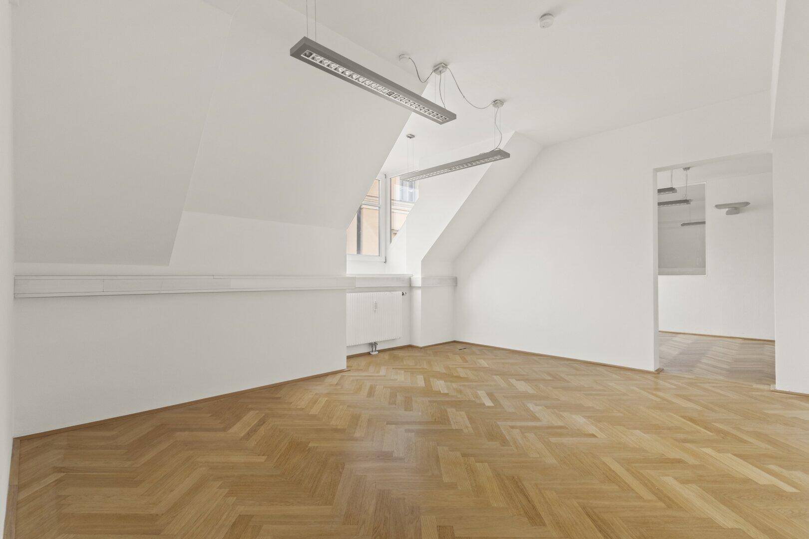 Geräumiger, heller Büroraum im Dachgeschoss mit schrägen Wänden, Fischgrätparkett und Blick in den Nebenraum.