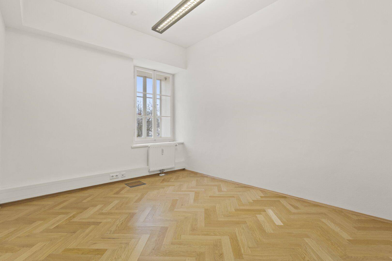 Kleinerer, heller Büroraum mit Fischgrätparkett und einem großen Fenster, das für natürliche Belichtung sorgt.