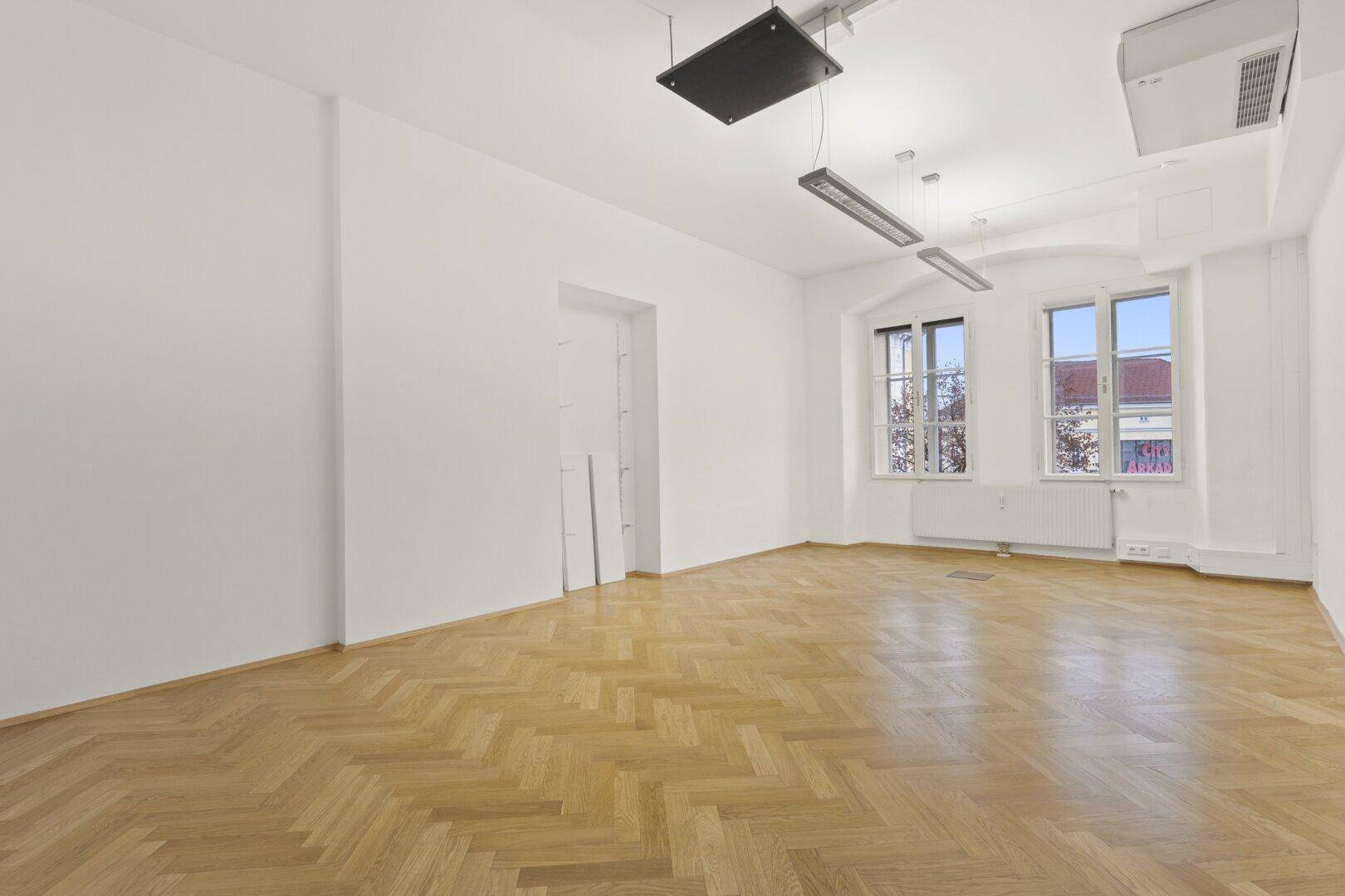 Heller Büroraum mit Fischgrätparkett, zwei Fenstern und einem offenen Durchgang, der zu weiteren Räumen führt.