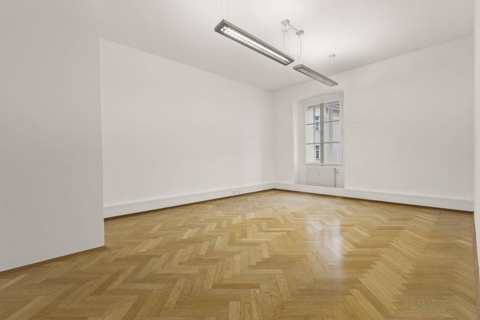Heller, leerer Raum mit Fischgrätparkett und einem Fenster, das für eine angenehme Arbeitsatmosphäre sorgt.