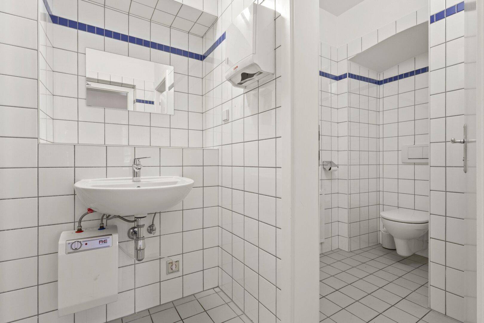 Helles Badezimmer mit weißen Fliesen, einem blauen Akzentstreifen und einem Waschbecken mit Toilette.
