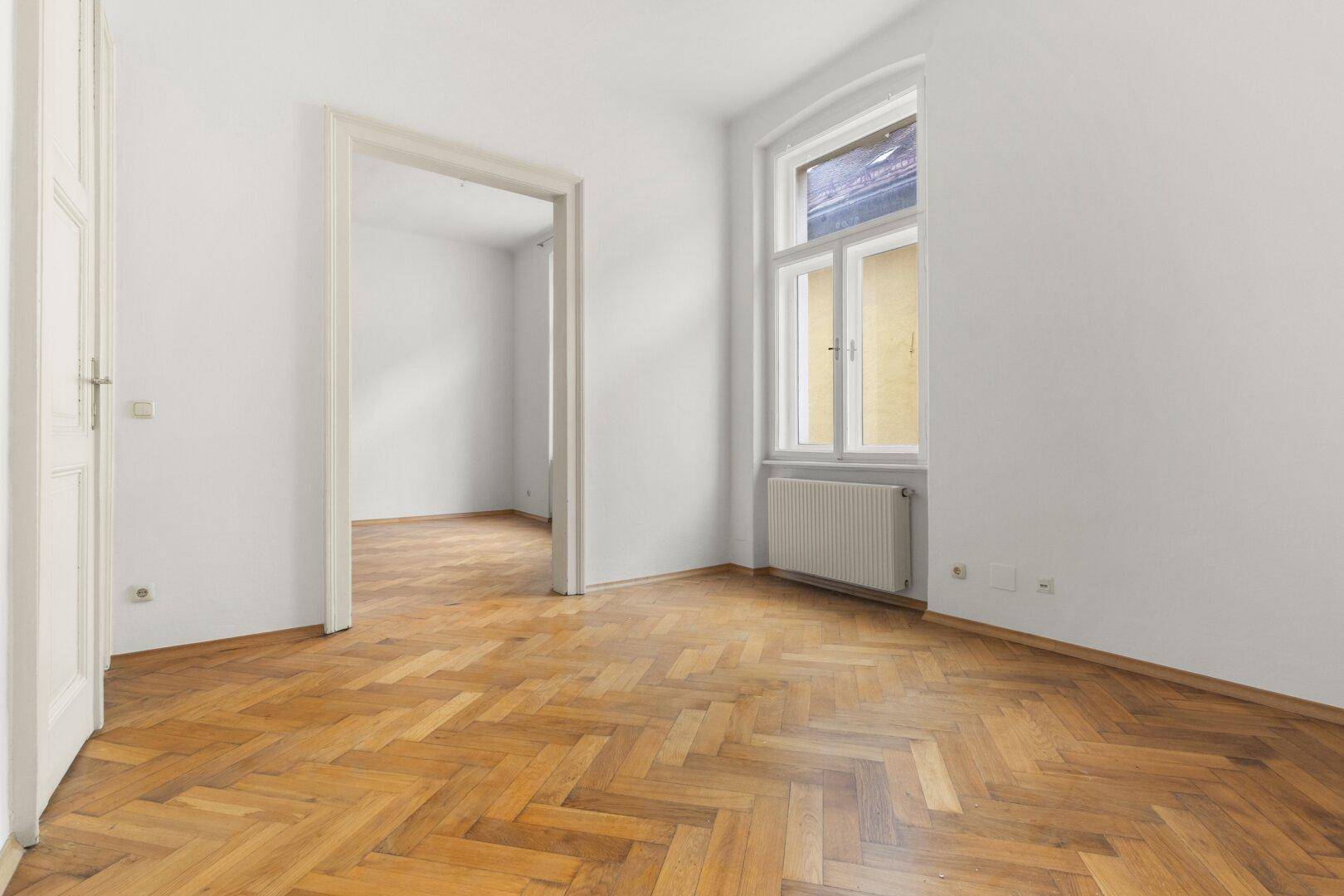 Freundlicher Raum mit Holzboden und Blick durch eine breite Tür in einen weiteren hellen Bereich.