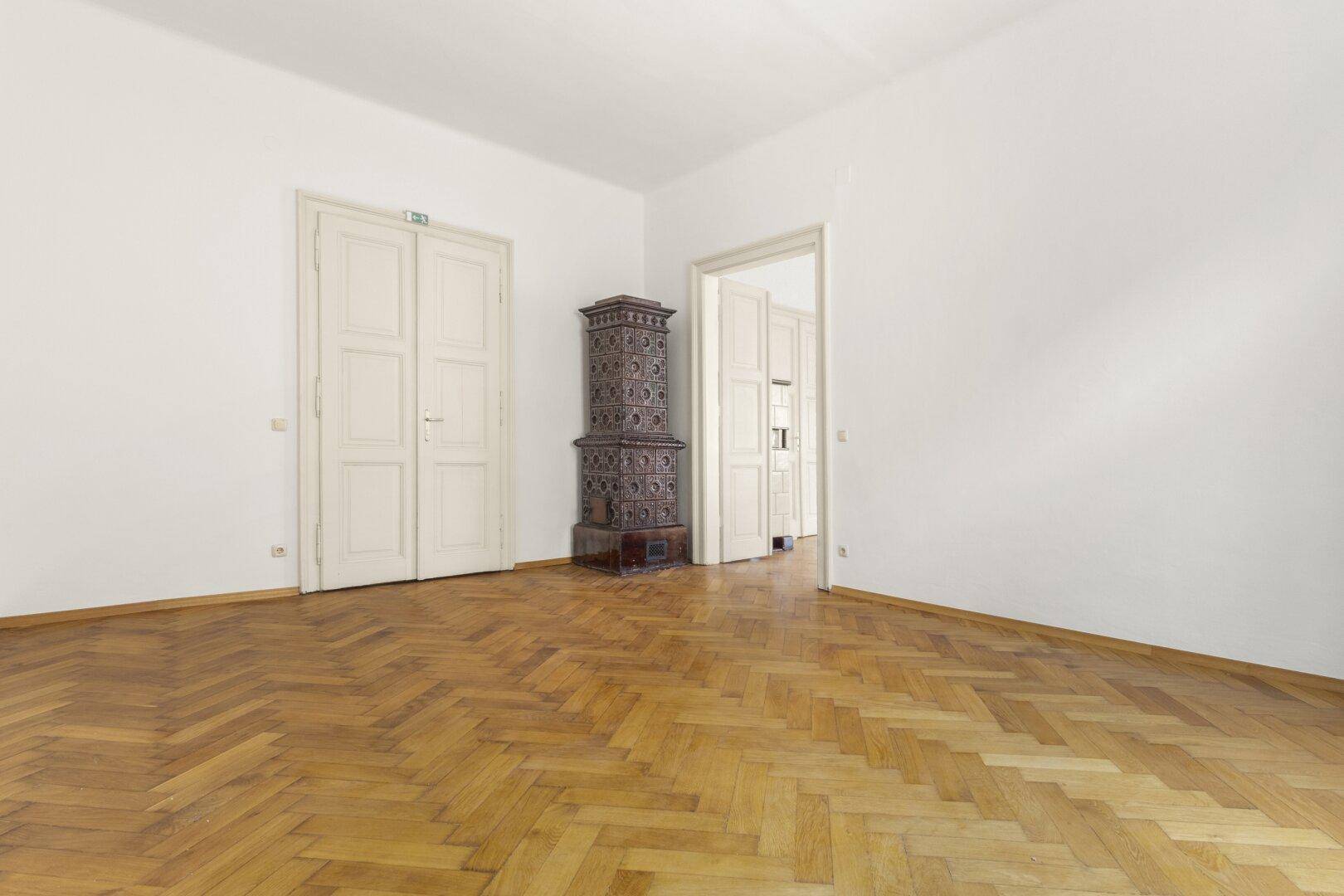 Heller Raum mit klassischem Fischgrätparkett, einem traditionellen Kachelofen und hohen Decken.