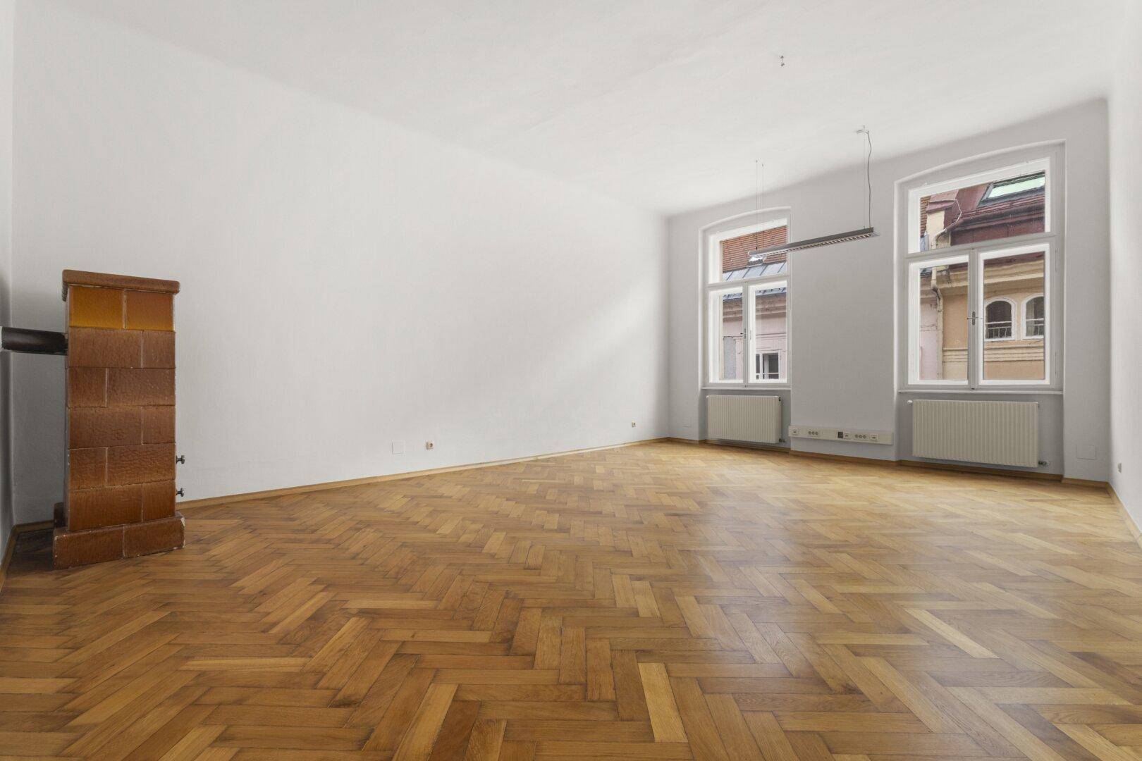 Großer, heller Raum mit Fischgrätparkett, hohen Fenstern und einem Kachelofen mit Stadtblick.